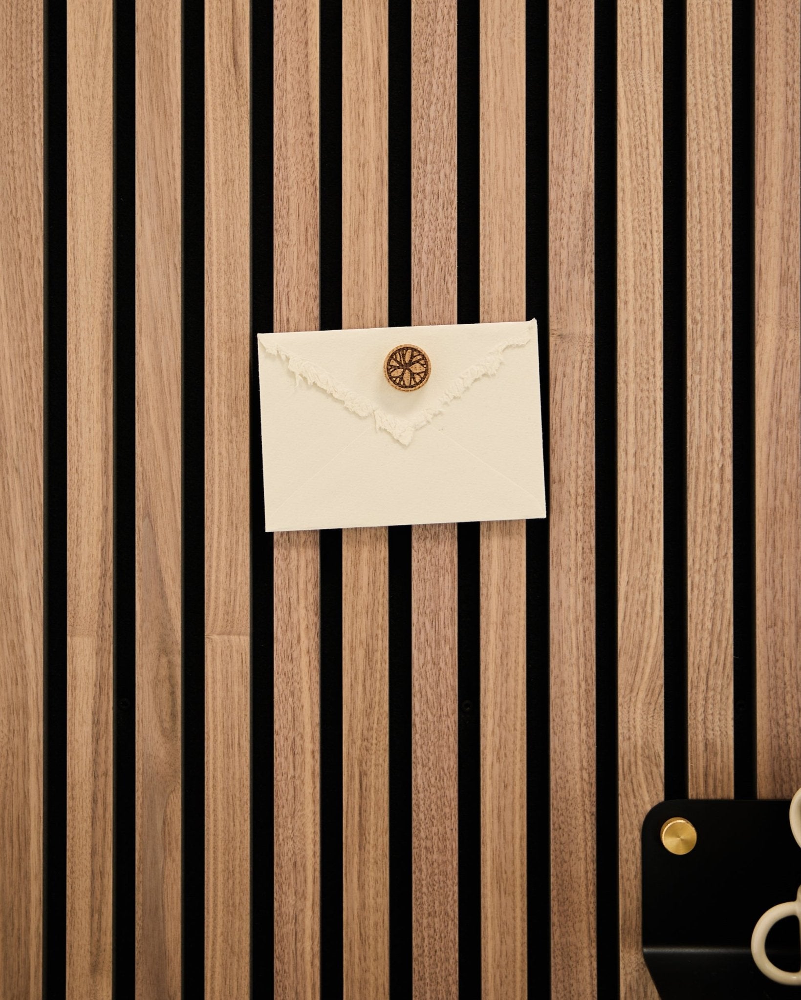 Una busta color crema con sigillo in ceralacca è fissata a un pannello slatwall acustico WoodUpp Rovere con il set di magneti per poster Norr in Natural Oak; forbici e vassoio nelle vicinanze.