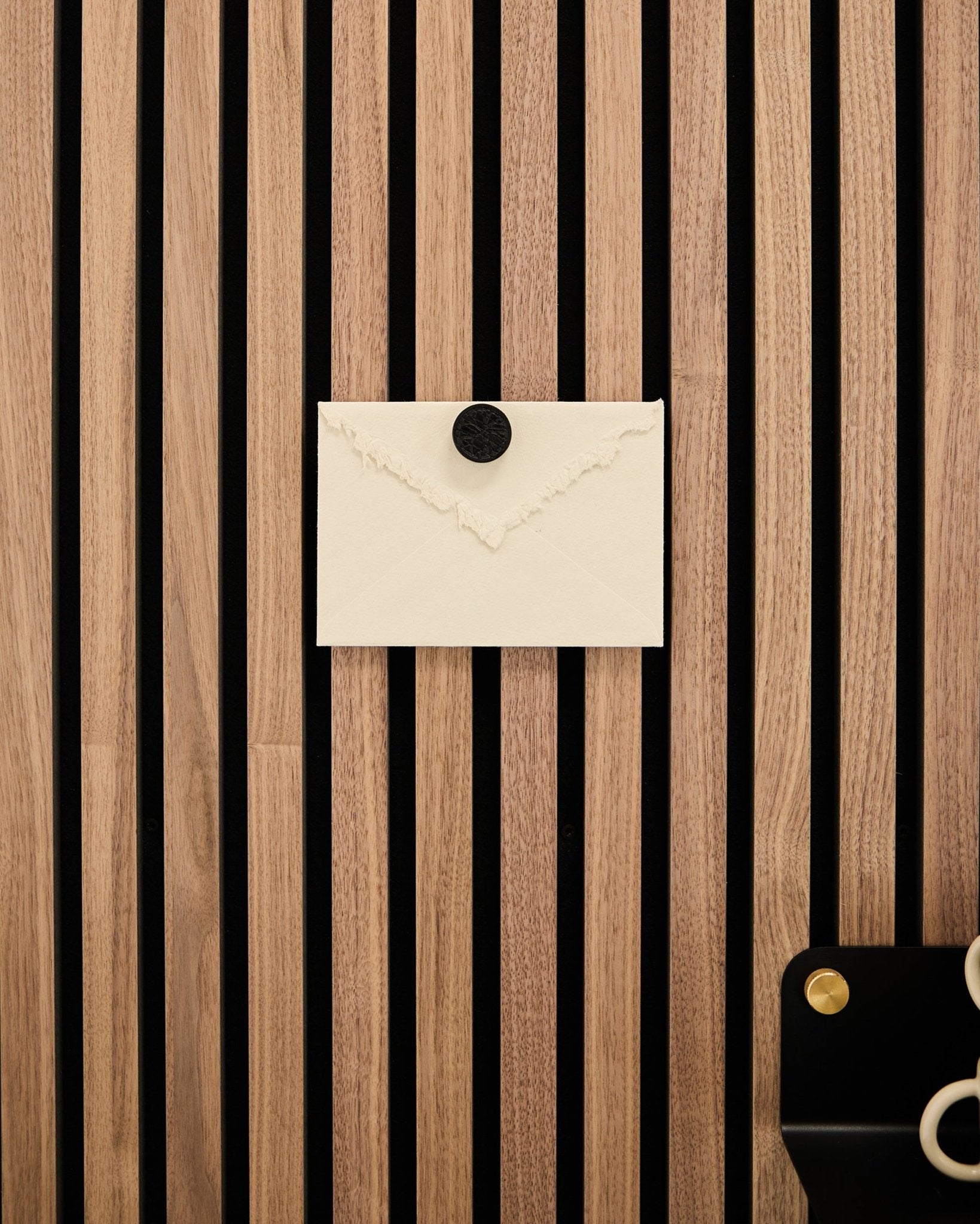 Una busta color crema è fissata a un pannello acustico a doghe WoodUpp Rovere a righe Pannello acustico con il set di magneti per poster Norr in Black Oak; in basso a destra ci sono delle forbici.