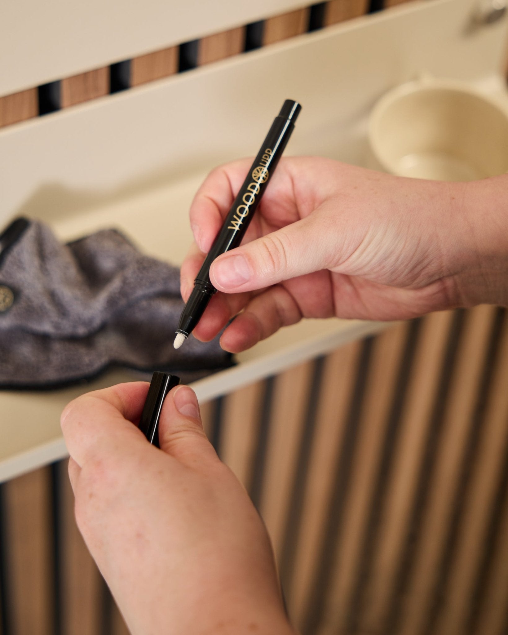 Una persona tiene in mano un pennarello WoodUpp Norr | Blackboard set aperto accanto a un panno in microfibra e a una tazza beige, prodotti pensati per l’uso con pannelli acustici in legno.