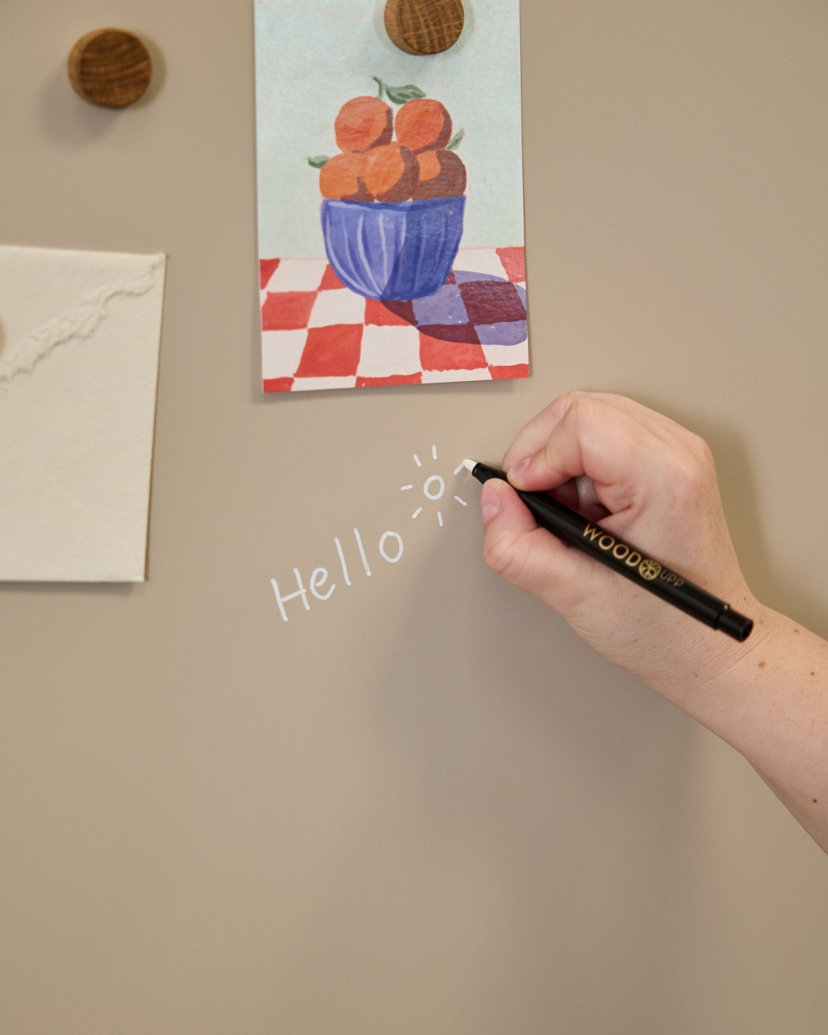 Una mano scrive hello con il disegno di un sole sulla WoodUpp Norr Blackboard, accanto ad alcune opere fissate con magneti rotondi su un Pannello acustico.