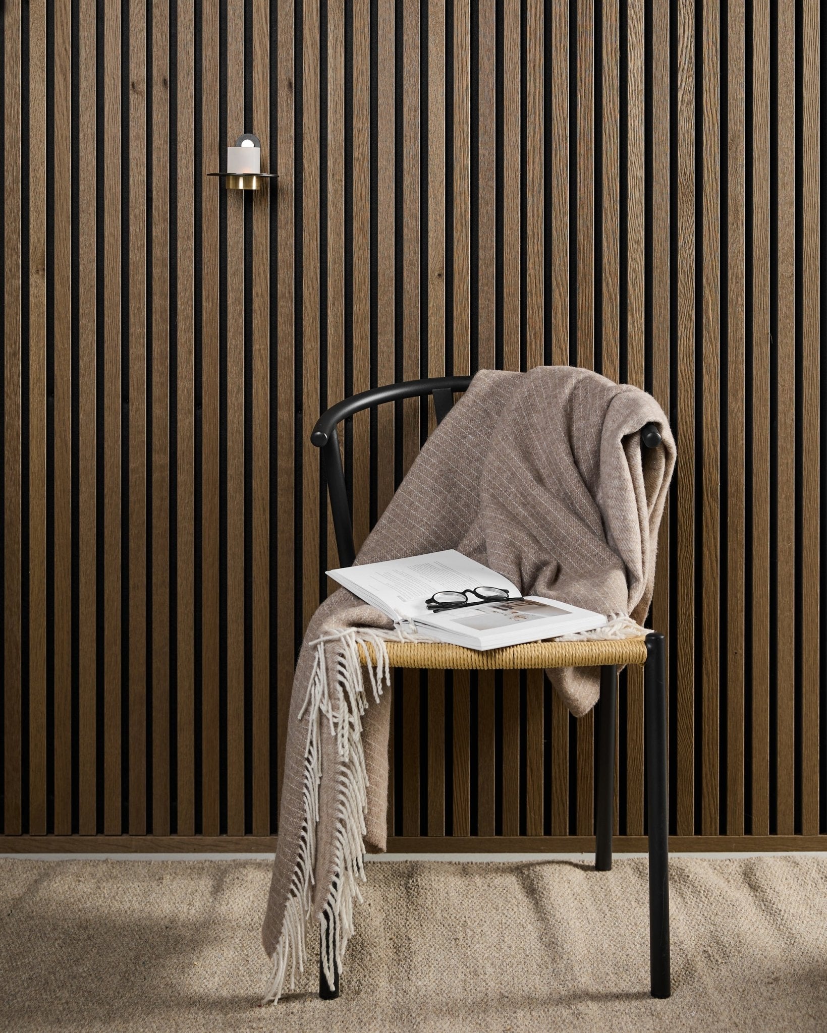 Una sedia moderna con un plaid beige, un libro aperto e un paio di occhiali è accanto a un pannello acustico in legno Akupanel e a un portacandela WoodUpp Norr black e brass.