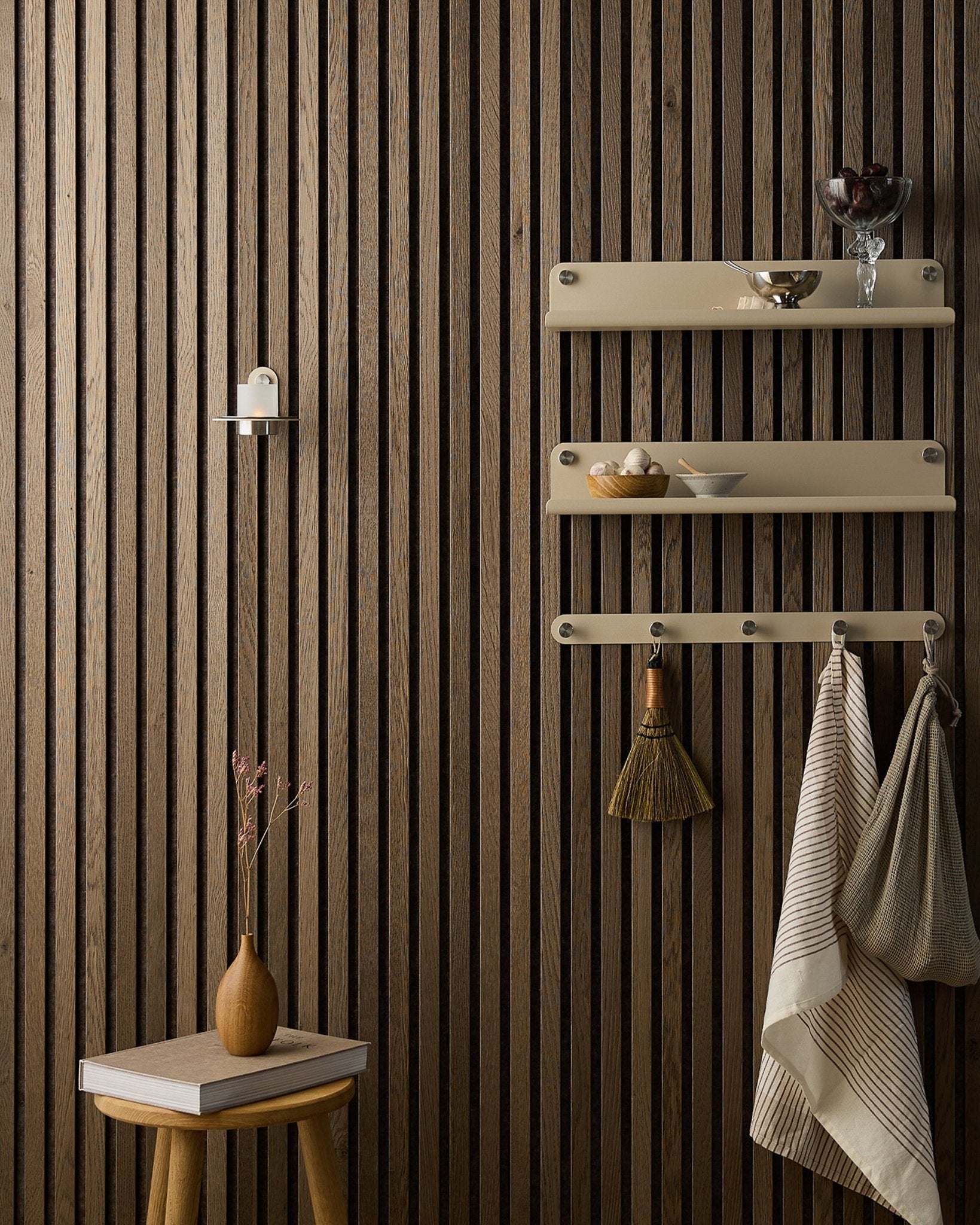 Uno sgabello in legno con fiori secchi è accanto a un Akupanel Pannello acustico di WoodUpp, vicino a mensole beige con piatti, una spazzola, asciugamani e un portacandele Norr.