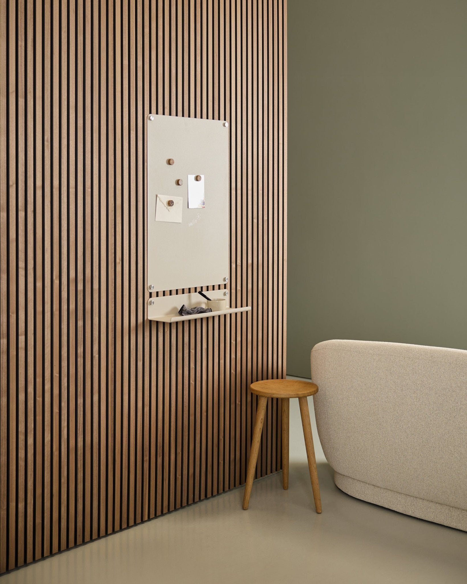 Interno minimalista con divano beige e sgabello in legno, con una lavagna magnetica WoodUpp Norr large beige montata su un pannello acustico in legno Nordic Akupanel vicino a una parete verde.