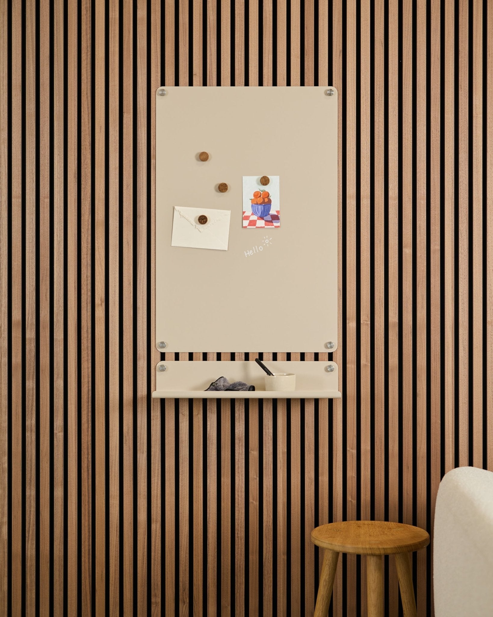 La lavagna magnetica Beige Norr di WoodUpp è montata sopra una mensola small su una parete con pannelli acustici in legno Akupanel, con uno sgabello e un divano bianco nelle vicinanze.