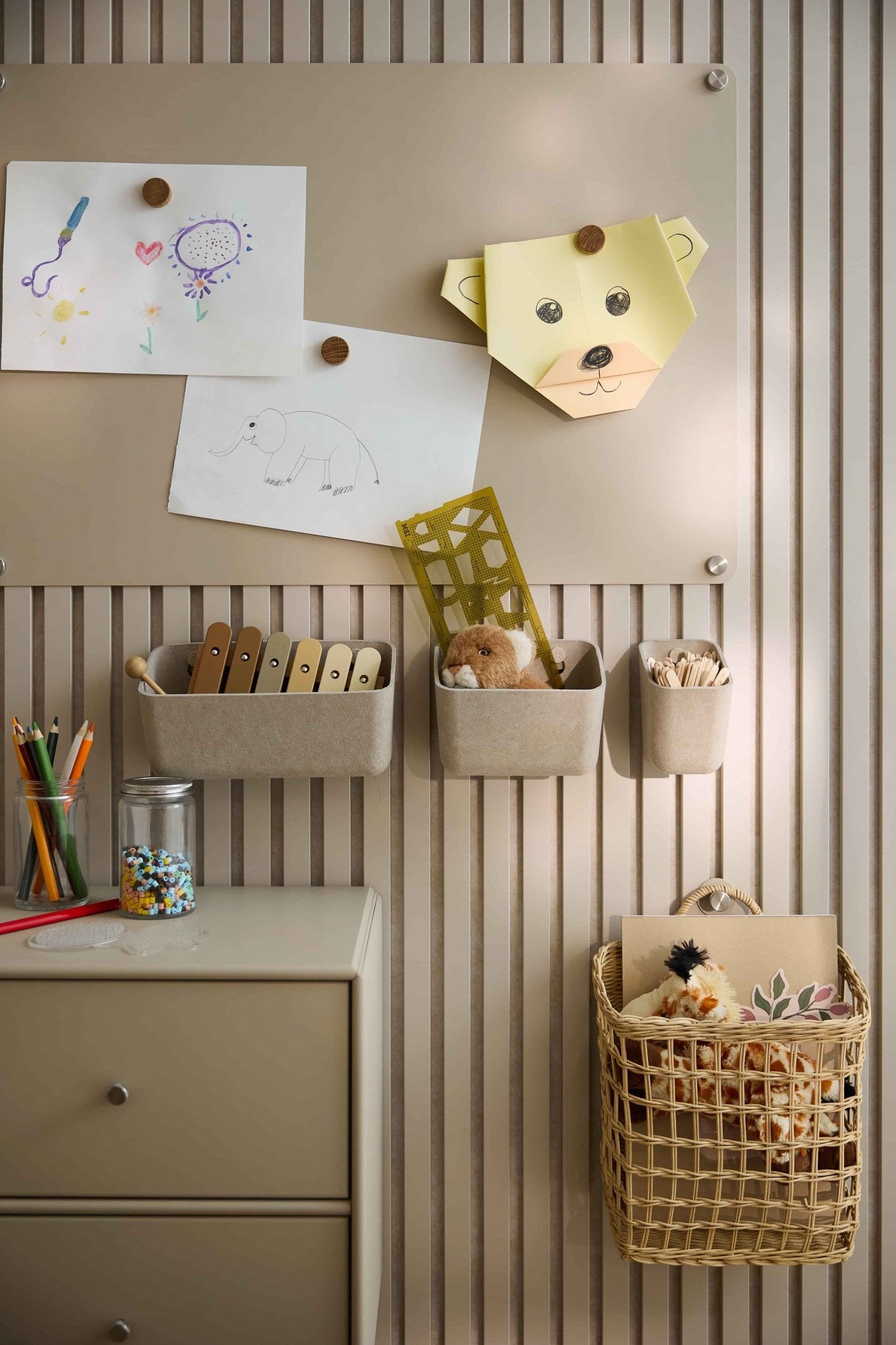 I disegni e i giocattoli dei bambini sono disposti su una parete beige rivestita con un pannello acustico WoodUpp, su cui sono collocati contenitori in feltro, cestini, un cassetto e un pannello forato.