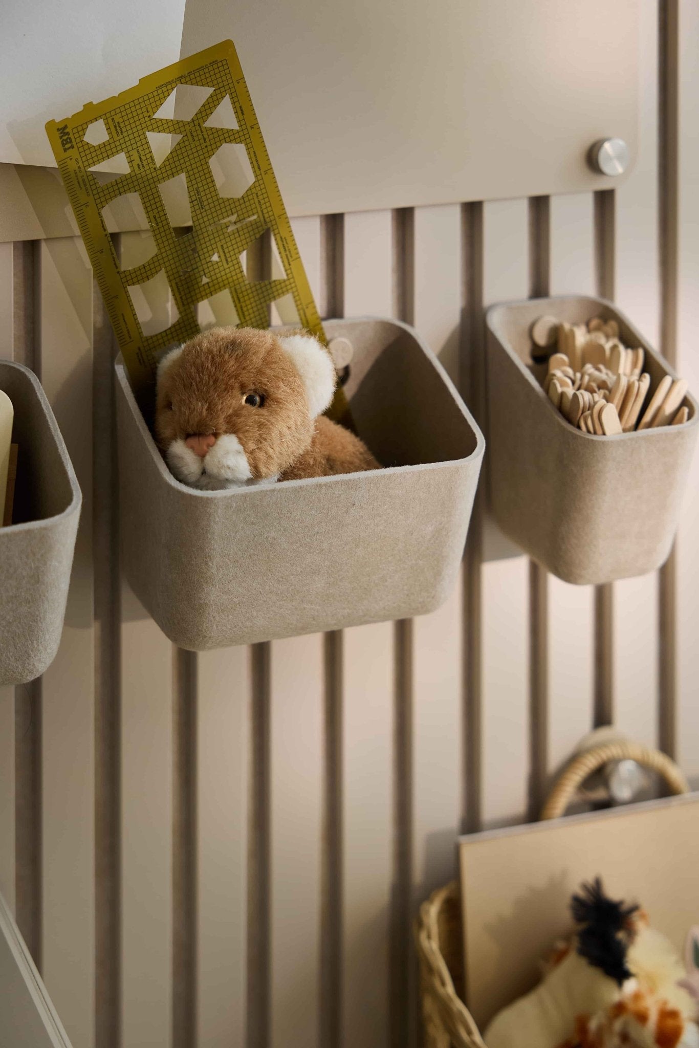 I contenitori in feltro WoodUpp Luva color beige contengono giocattoli, un righello giallo e bastoncini di legno, consentendo di riporre il tutto in modo ordinato su un pannello acustico a doghe a strisce.