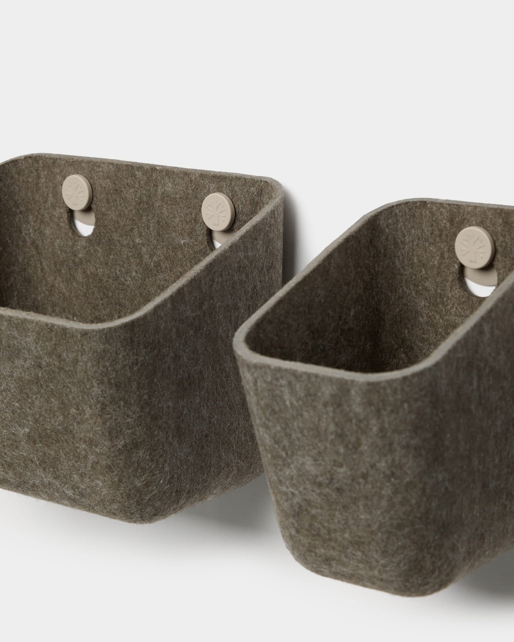 Due contenitori in feltro Luva, di colore grigio scuro caldo, realizzati da WoodUpp, sono appesi a ganci beige fissati a un pannello acustico, creando una soluzione minimalista per riporre oggetti a parete con angoli arrotondati.