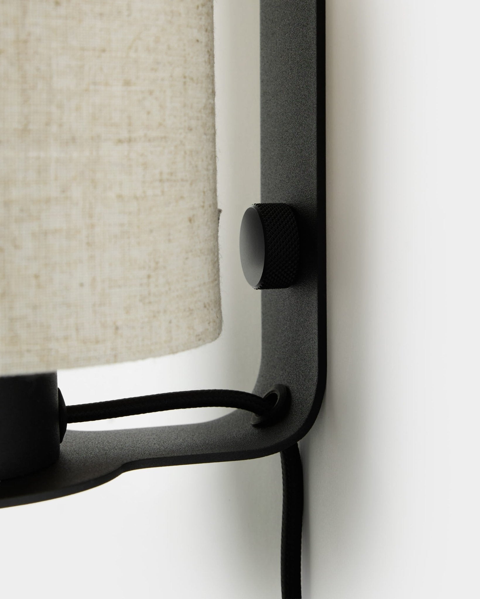Primo piano della lampada da parete Feya di WoodUpp in black e beige, con pomello testurizzato, paralume in lino-cotone e cavo a vista, montata su una parete bianca.
