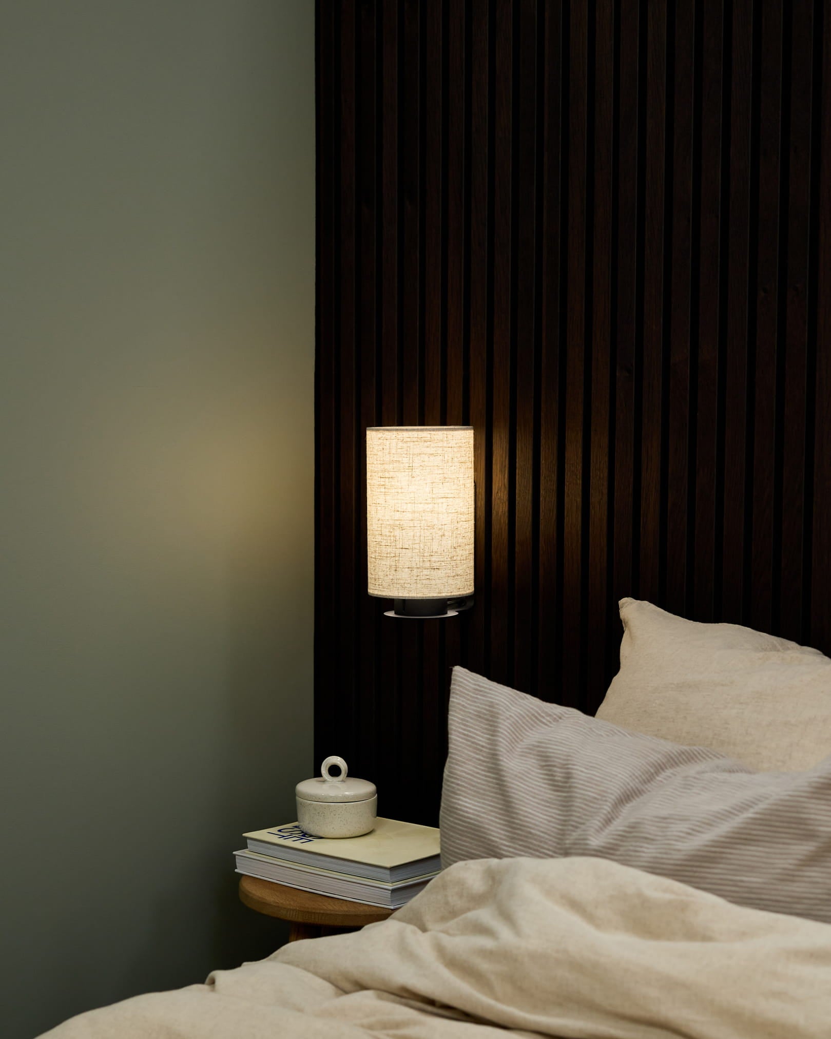 Angolo camera da letto con lampada da parete Feya di WoodUpp in Black/Beige, biancheria da letto beige e comodino con libri e una tazza in ceramica con coperchio.