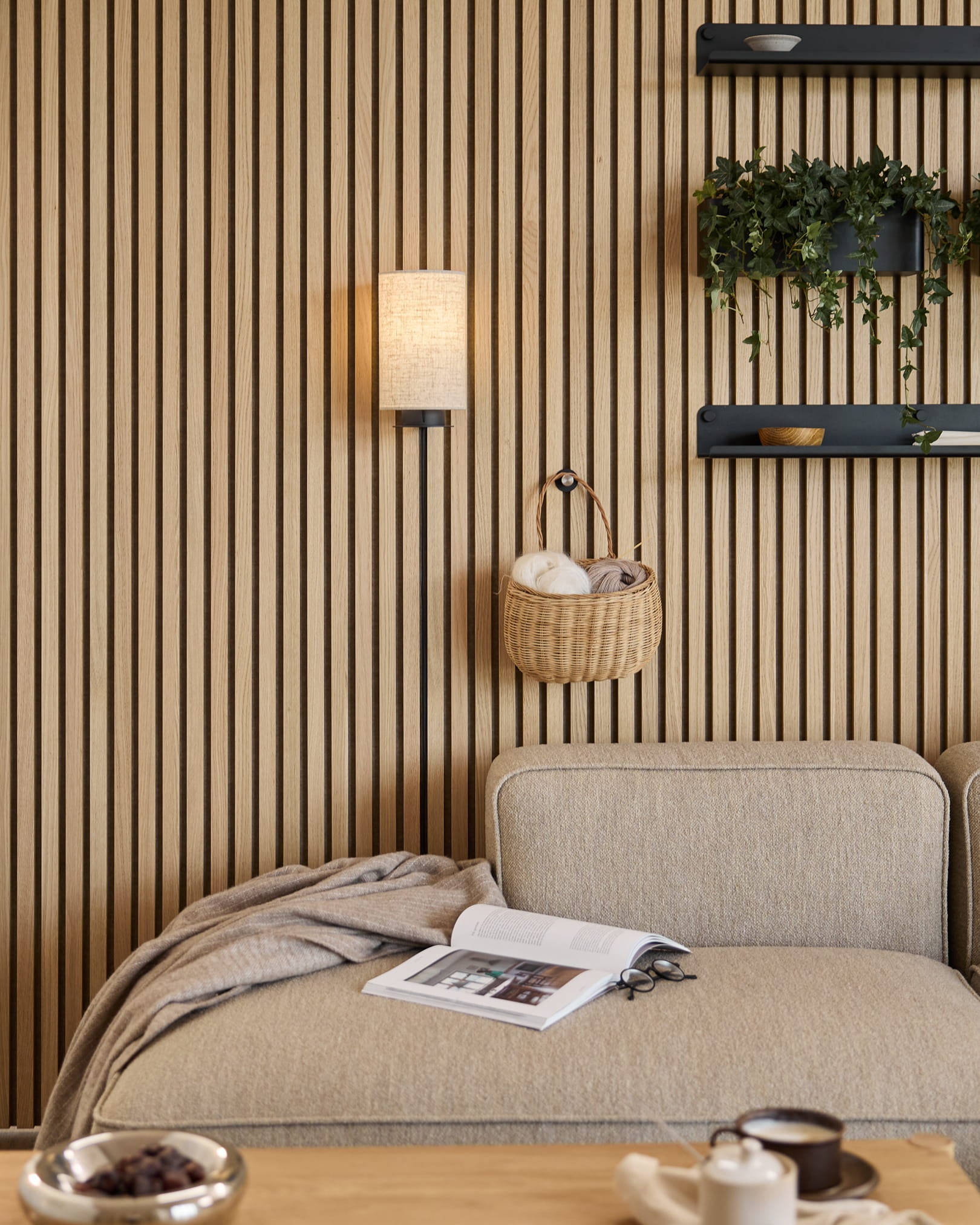 Divano Beige con coperta e libro accanto a una parete acustica a doghe WoodUpp Akupanel, mensole con piante e oggetti decorativi, e una lampada da parete Feya in Black/Beige.