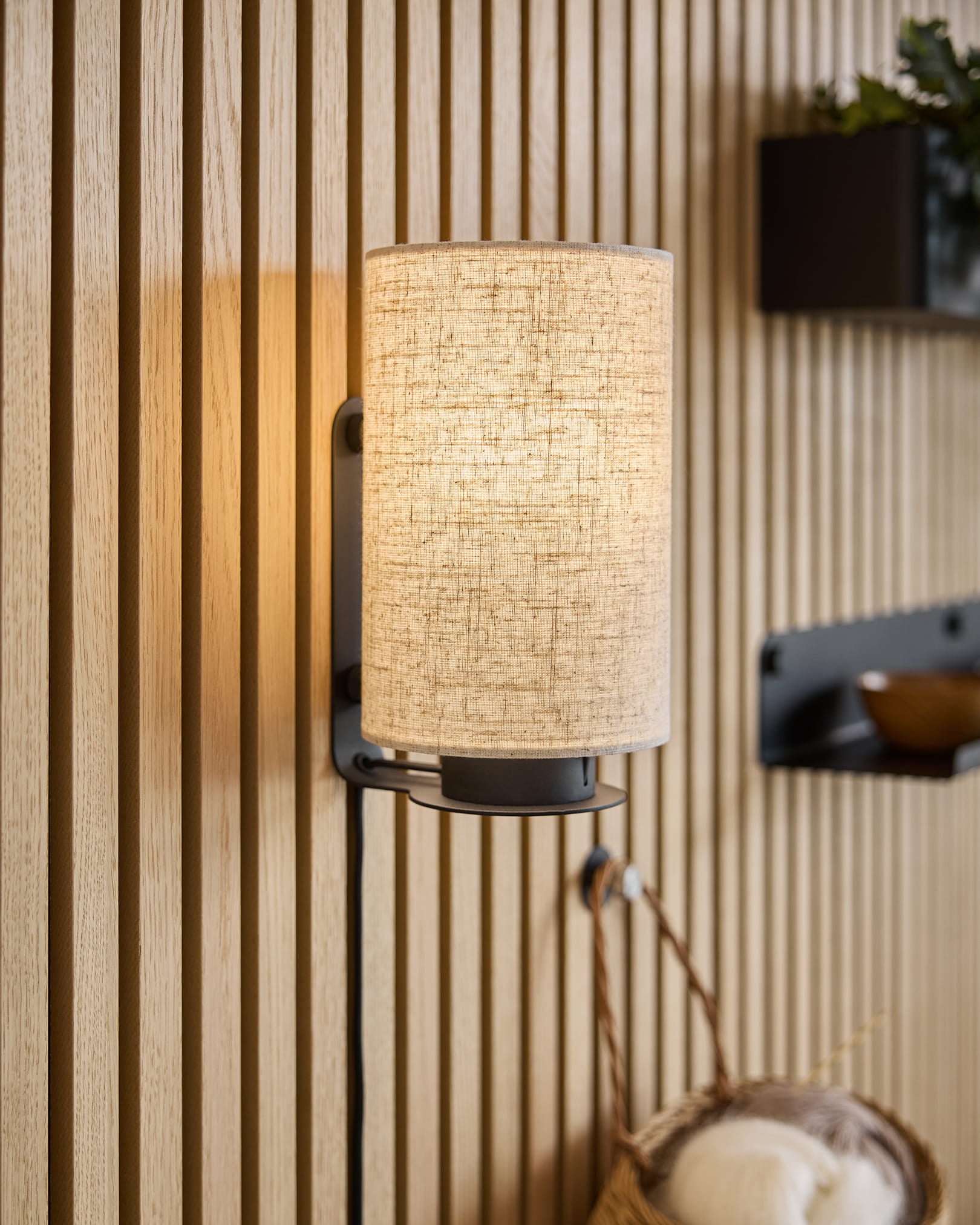 Una moderna applique con paralume in tessuto proietta una luce calda sui pannelli in legno acustico Akupanel, completati da mensole minimaliste e da un cesto intrecciato.