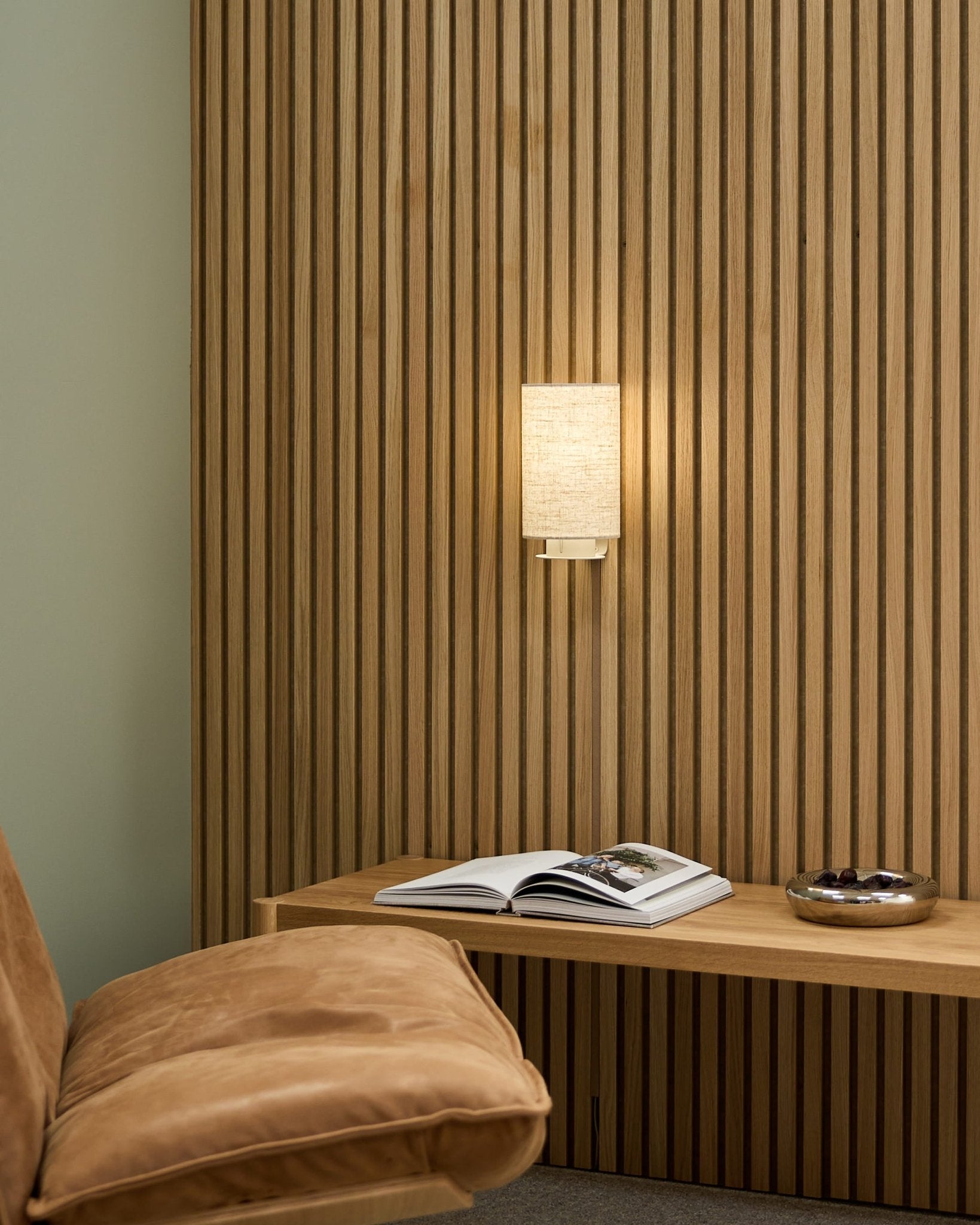 Stanza nordica minimalista con una sedia color cuoio, parete acustica a doghe WoodUpp Akupanel, Feya Wall Lamp in Beige/Beige, un libro aperto su una panca e una ciotola.