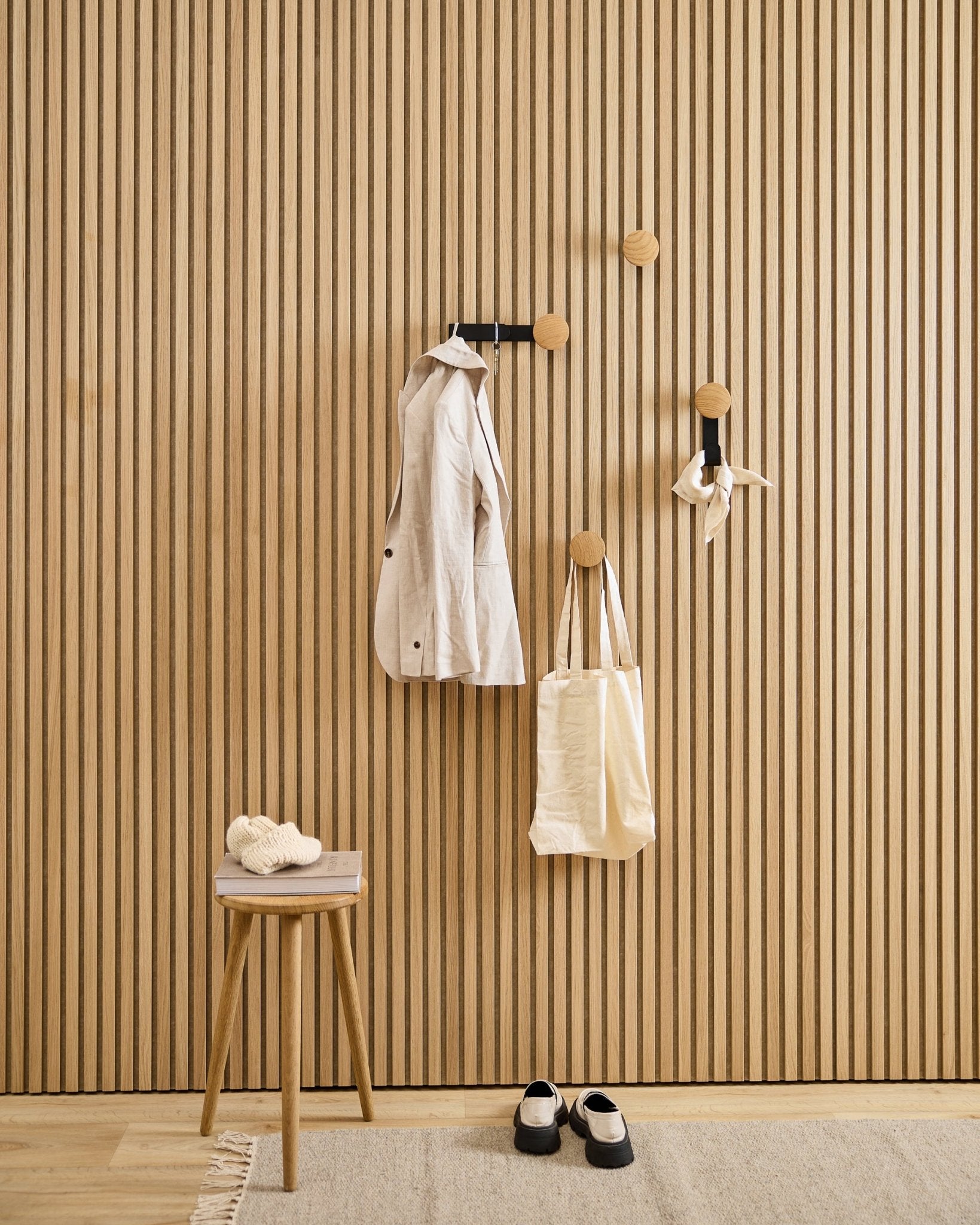 Ingresso minimalista con appendiabiti WoodUpp Linn Vertical in natural oak, con un cappotto beige, una tote bag, una sciarpa, uno sgabello con sopra delle pantofole e scarpe black e bianche.