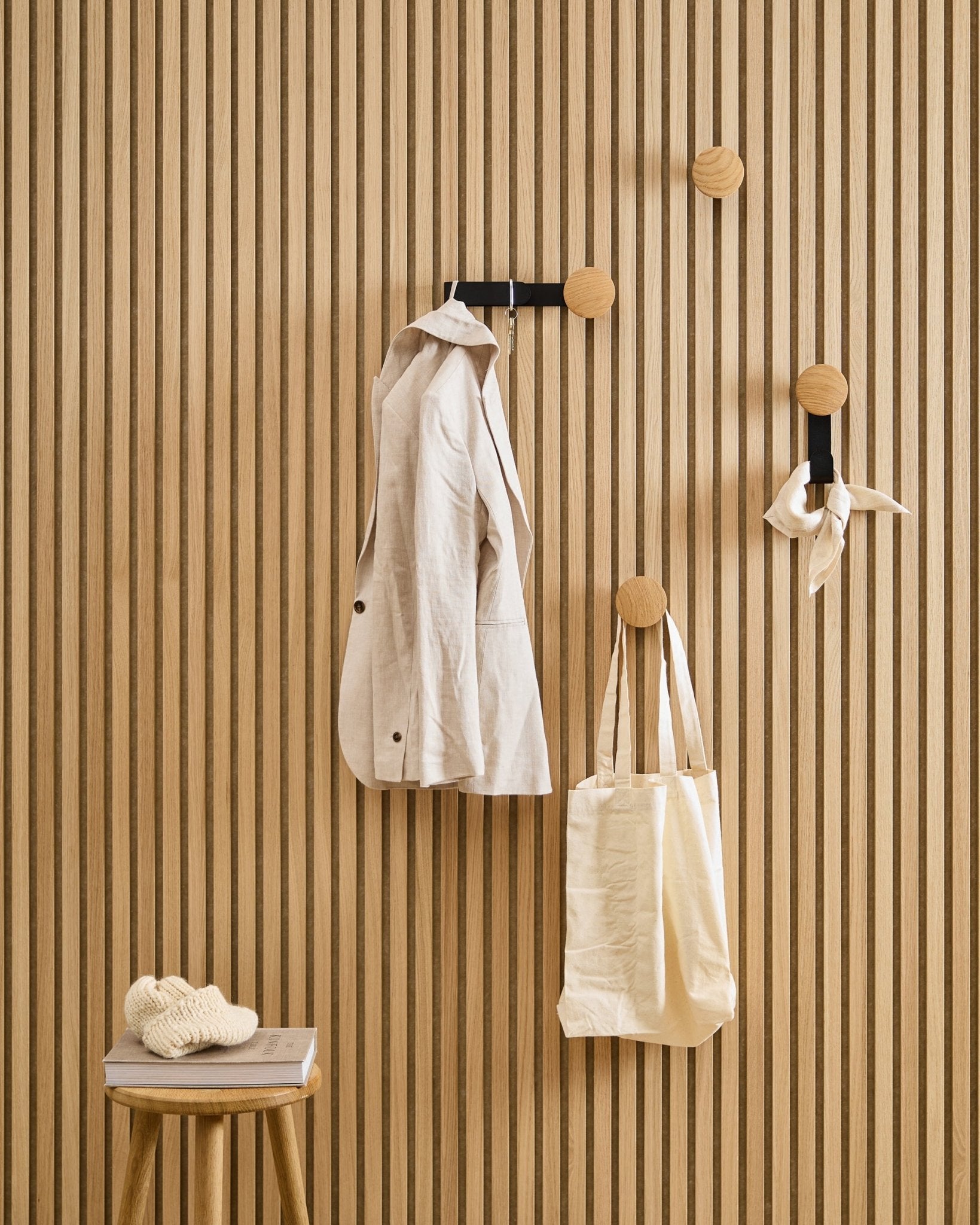 Una giacca beige, una tote bag e una sciarpa sono appese all’appendiabiti WoodUpp Linn Horizontal in Natural Oak; su uno sgabello accanto sono appoggiati un libro e un capo lavorato a maglia.