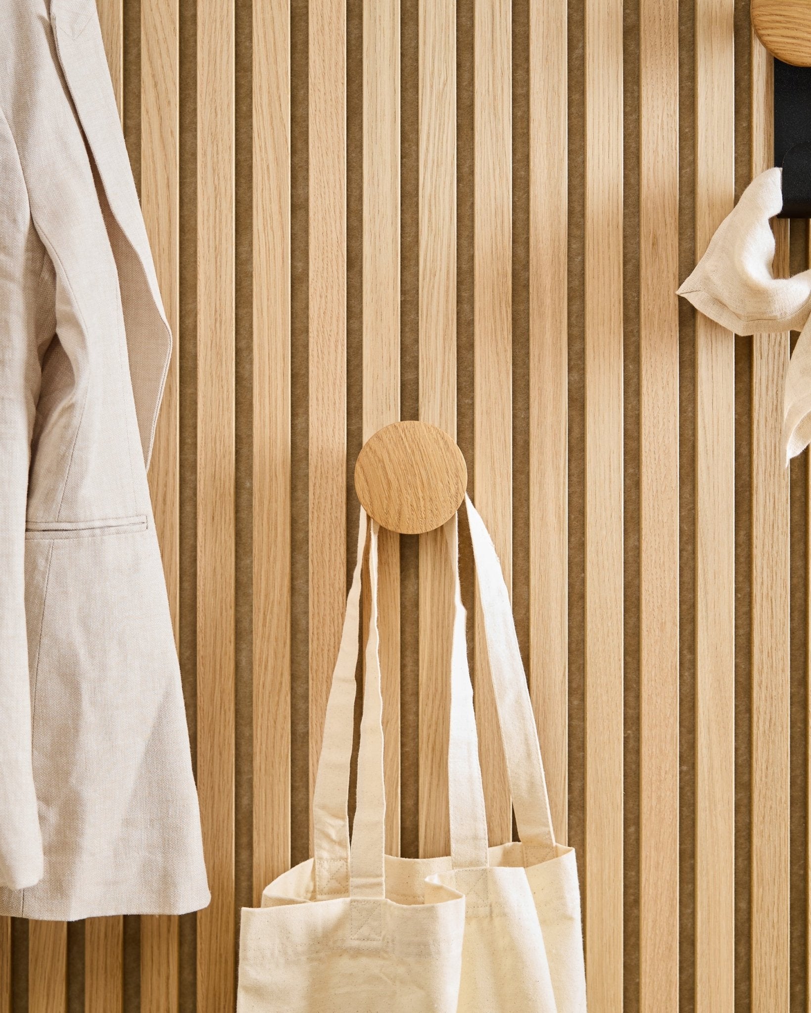Un cappotto beige e una tote bag sono appesi al Linn I Single Hanger in Natural Oak di WoodUpp, montato su un pannello acustico in legno a doghe vertical Akupanel.