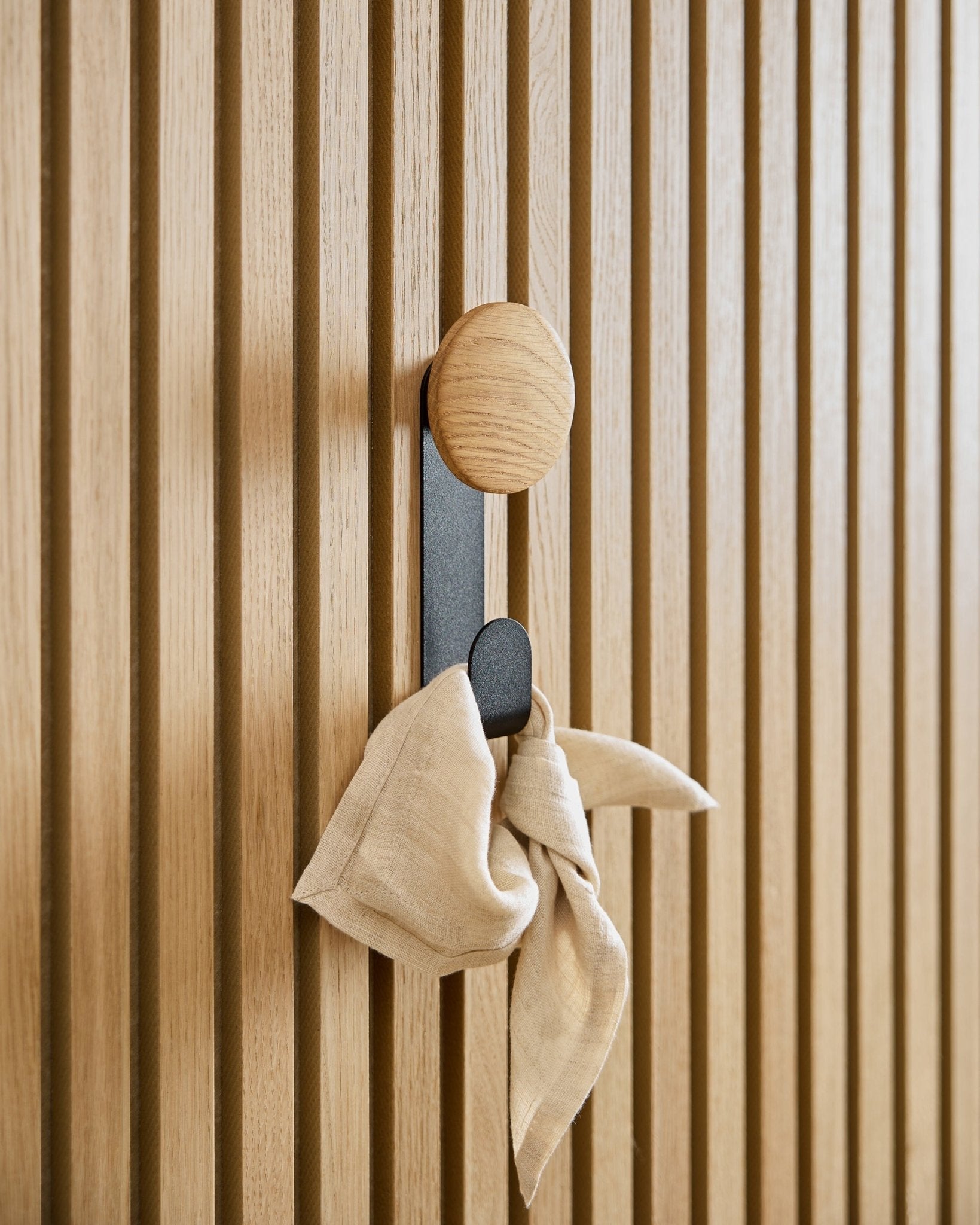 Un panno in beige è appeso all’appendiabiti Linn Vertical in Natural Oak di WoodUpp, montato su un pannello acustico a listelli vertical Akupanel.