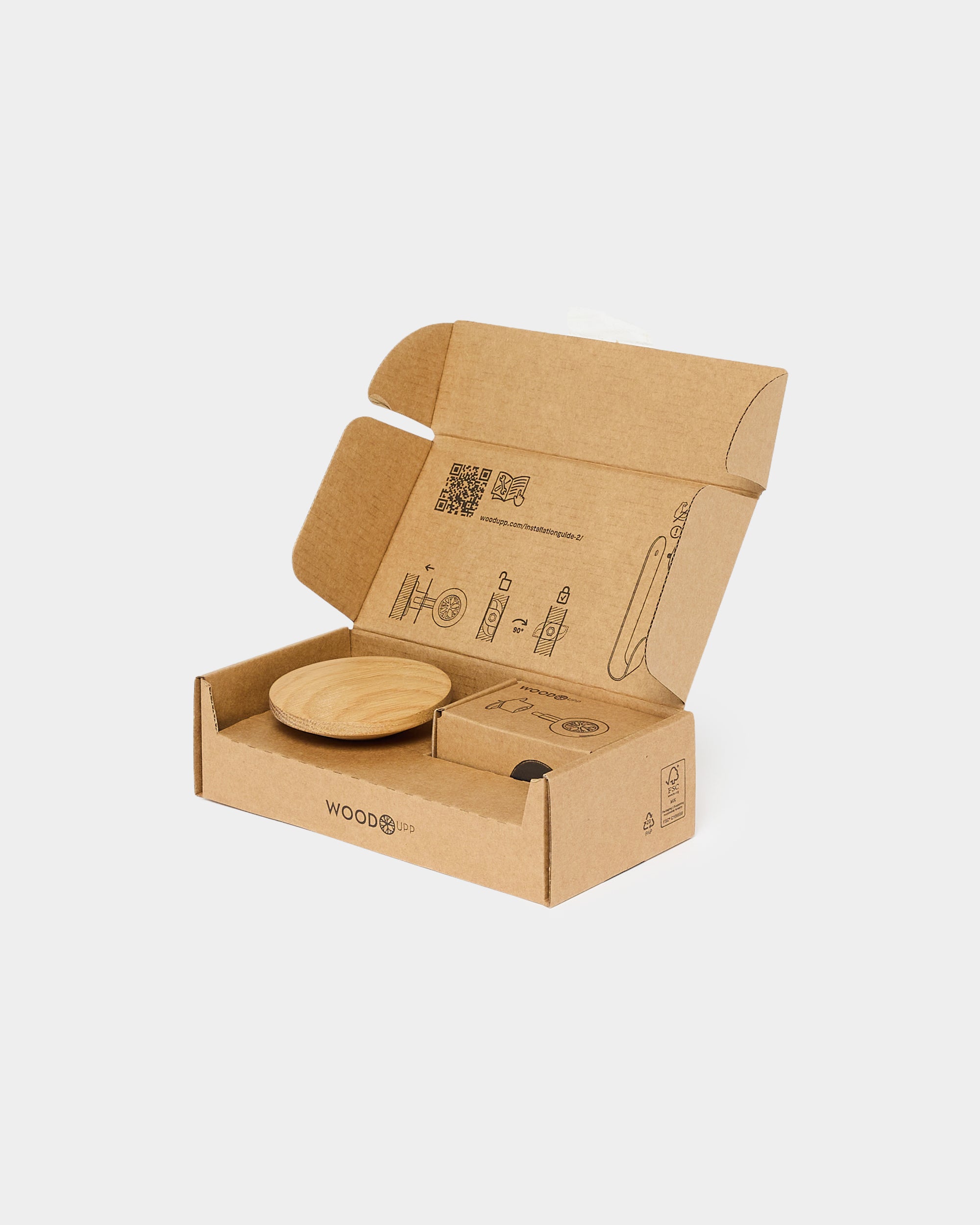 A circular wooden accessory and printed assembly instructions from WoodUpp are placed inside an open cardboard box, prepared for Akupanel installation.