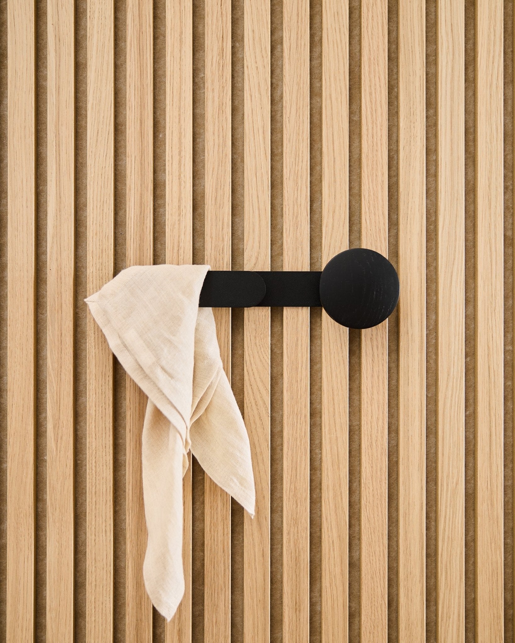 Un panno beige è appoggiato sul gancio WoodUpp Linn Horizontal in Black Oak, fissato a un pannello acustico a listelli vertical.