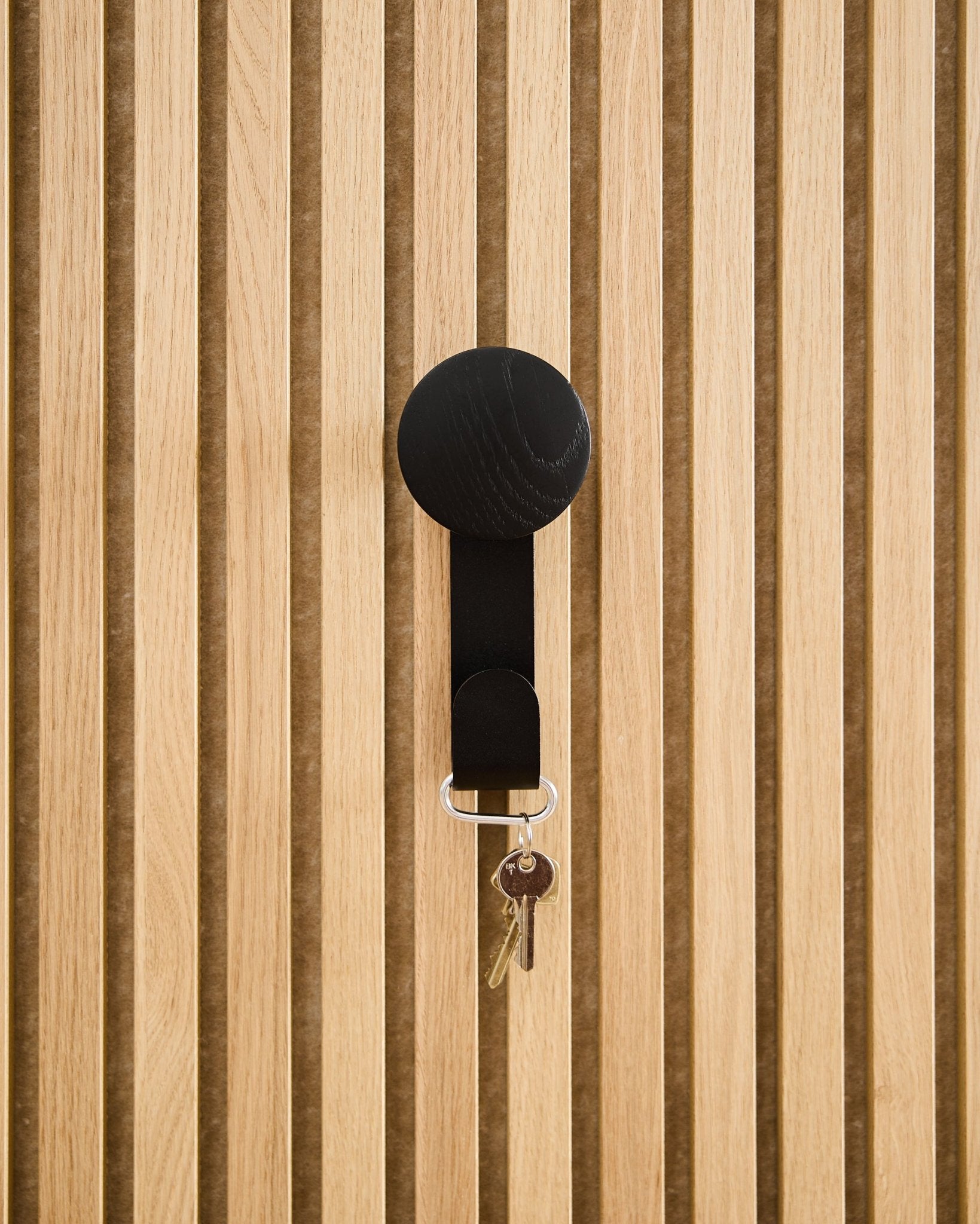 Un appendiabiti Linn Vertical in Black Oak di WoodUpp sostiene un portachiavi con due chiavi e un tag rotondo, montato su un pannello acustico in legno Akupanel dalla superficie testurizzata.