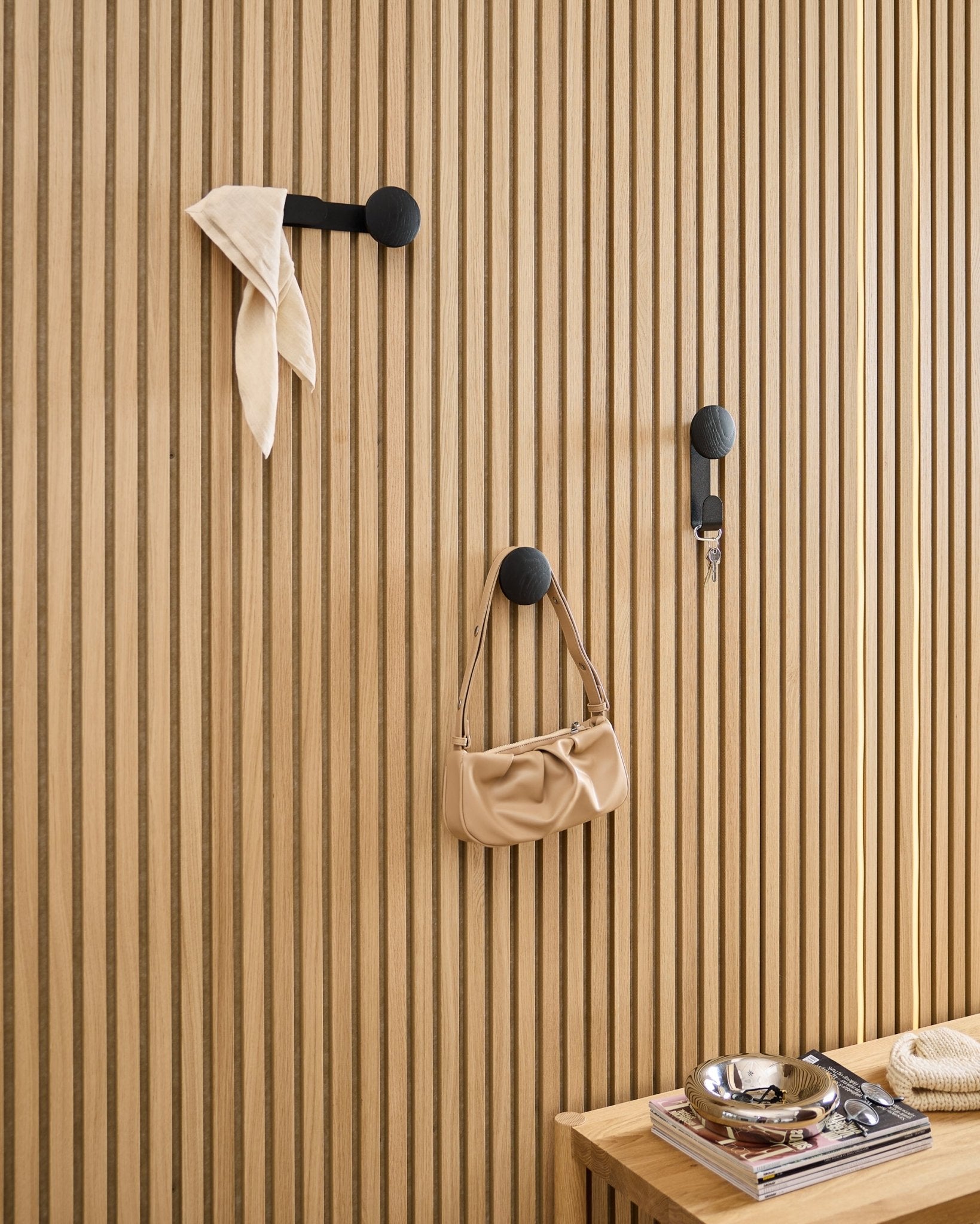 Il/La Linn Single Hanger di WoodUpp in Black Oak sostiene una sciarpa, una borsa beige e delle chiavi su un pannello acustico in legno Akupanel black; nelle vicinanze una panca con libri e un piatto in metallo.