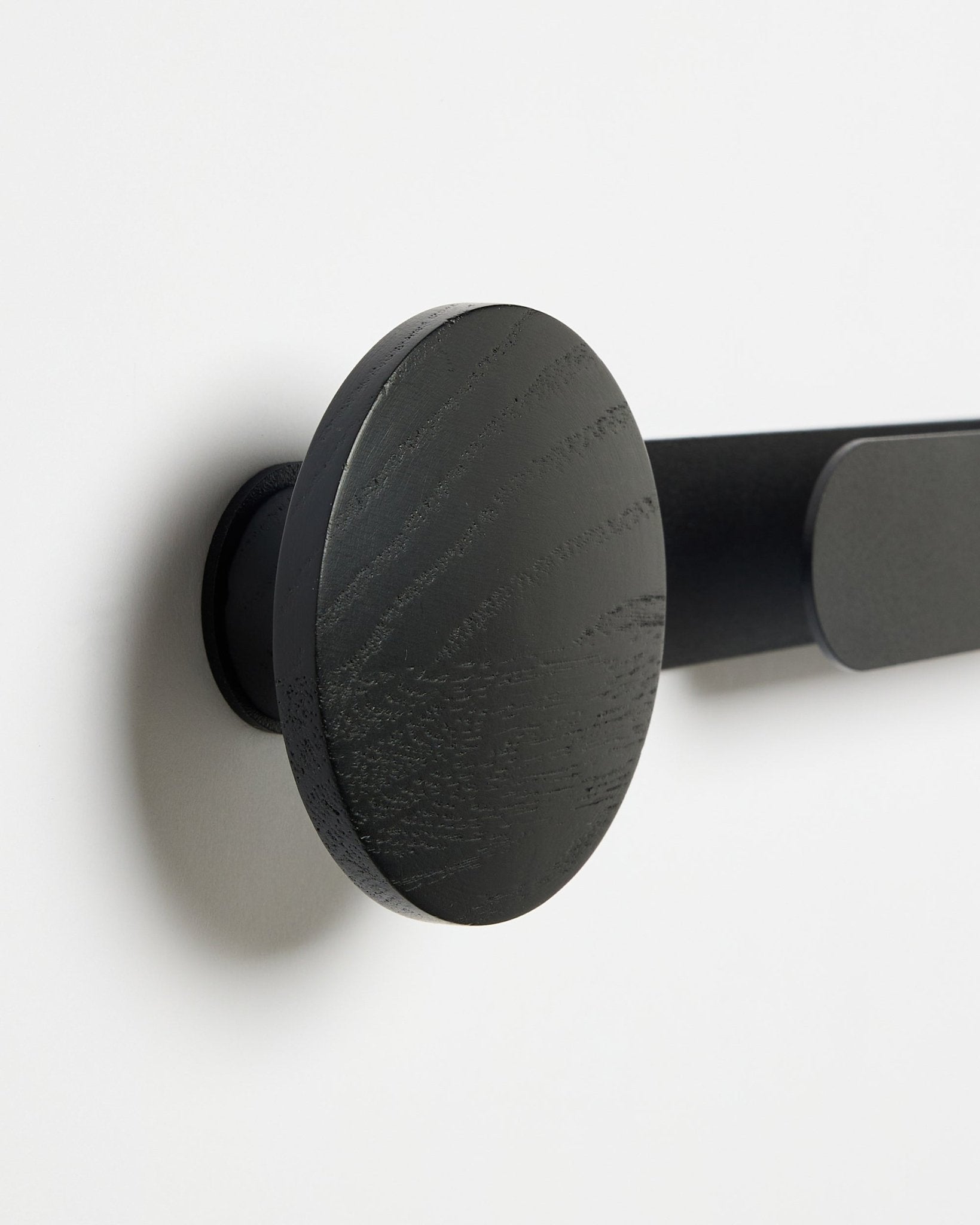 Primo piano del gancio WoodUpp Linn | Horizontal in Black Oak, finitura opaca black, montato su una parete bianca; accessorio per pannelli acustici in legno.