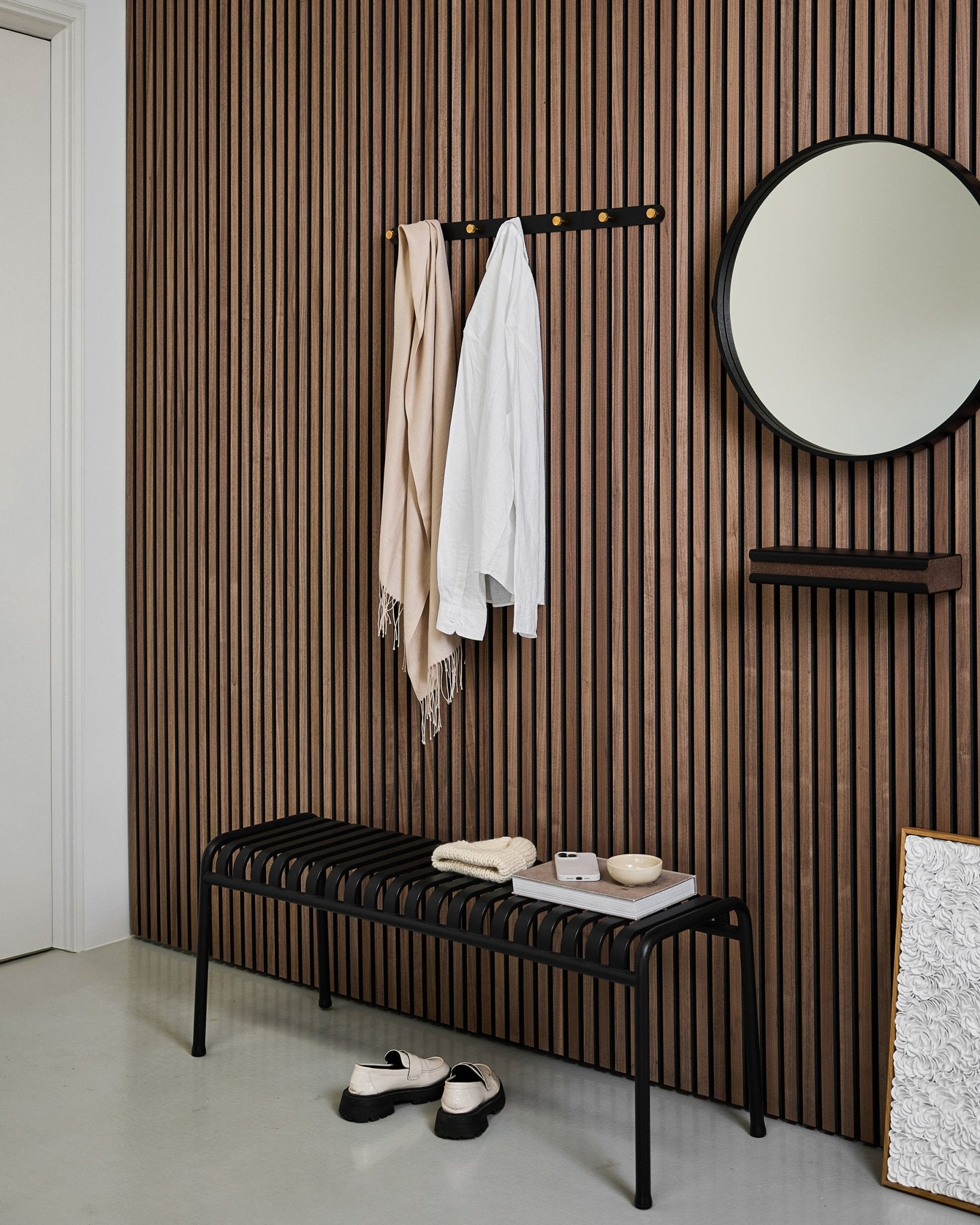 Ingresso minimalista con una parete acustica a listelli WoodUpp Akupanel, appendiabiti Norr Coat Hook Rack (Black/Brass, X7), panca, specchio e arredi moderni dai toni neutri.