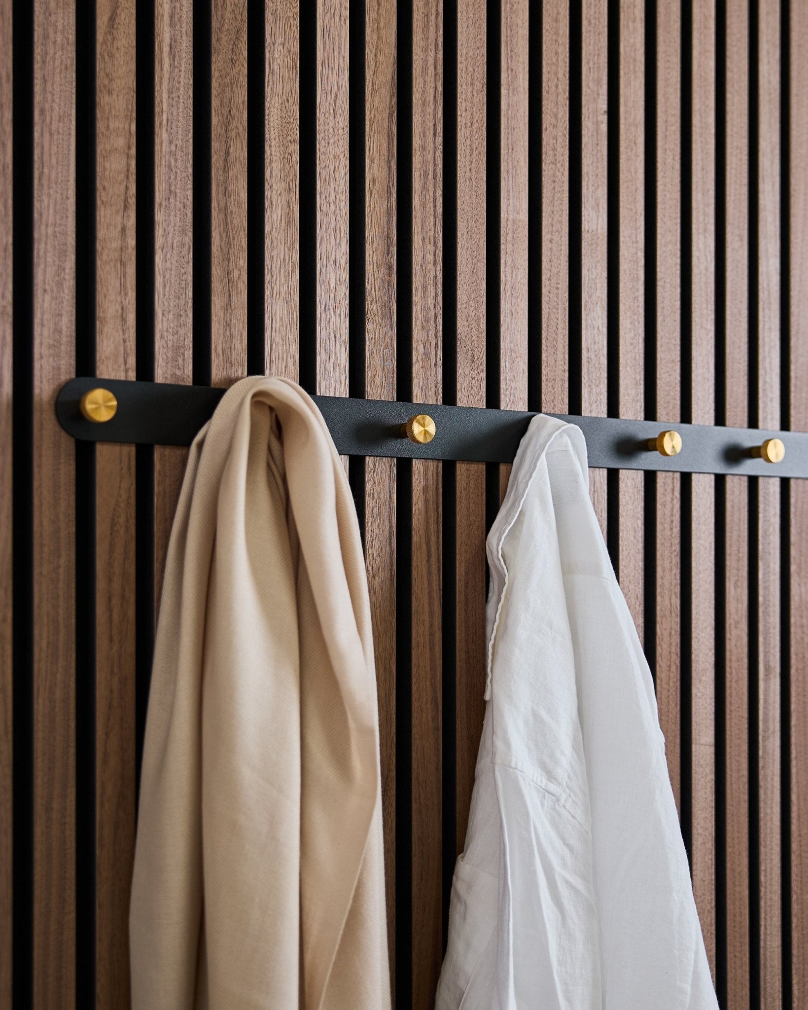 Il Coat Hook Rack WoodUpp Norr in black/brass (X7), con una sciarpa beige e una camicia bianca, è appeso a un pannello acustico nordico in legno a listelli.