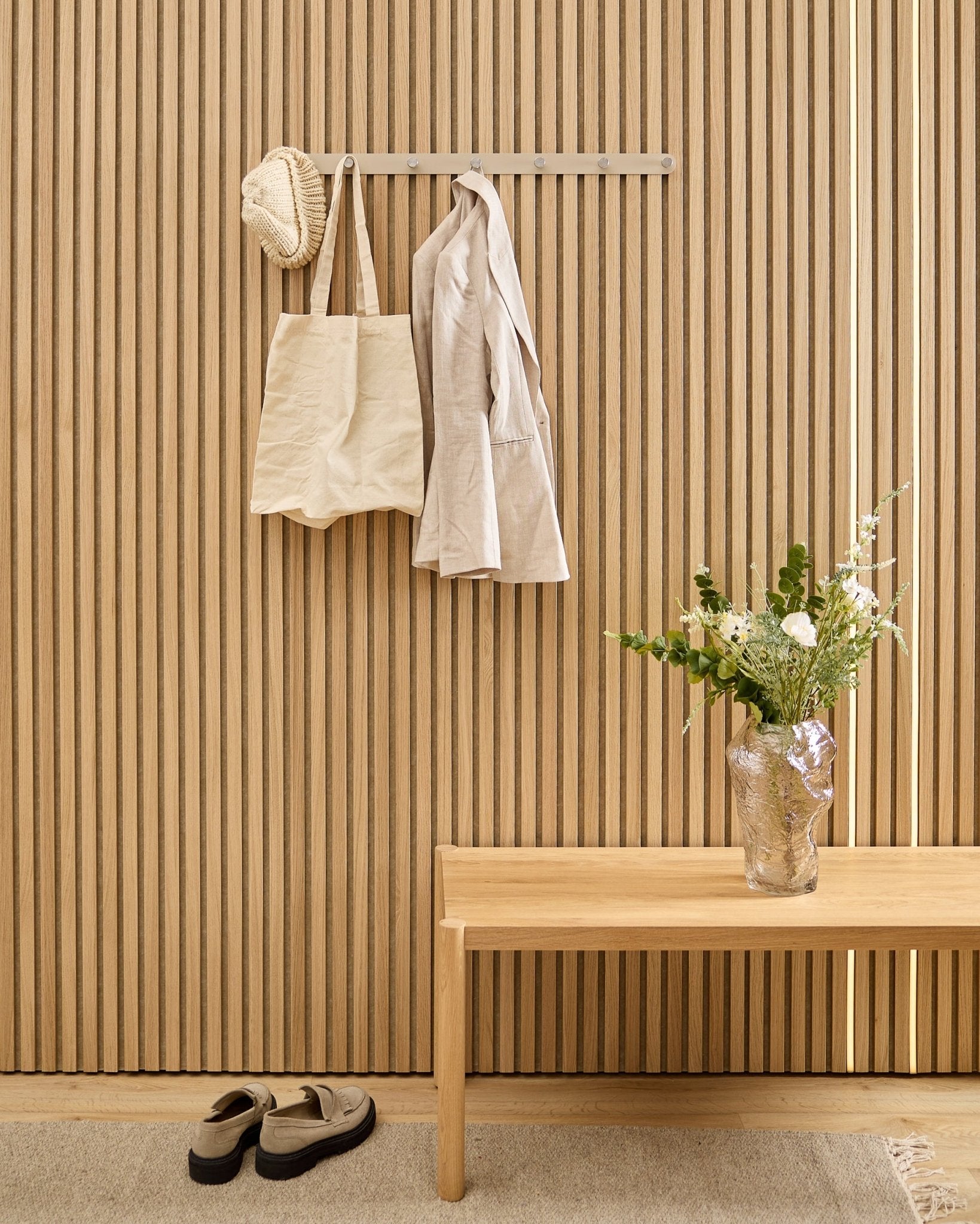 Una borsa beige, un cappello e una camicia sono appesi all’appendiabiti a ganci WoodUpp Norr sopra le scarpe, accanto a una panca e a un vaso vicino al pannello acustico a doghe rigate.