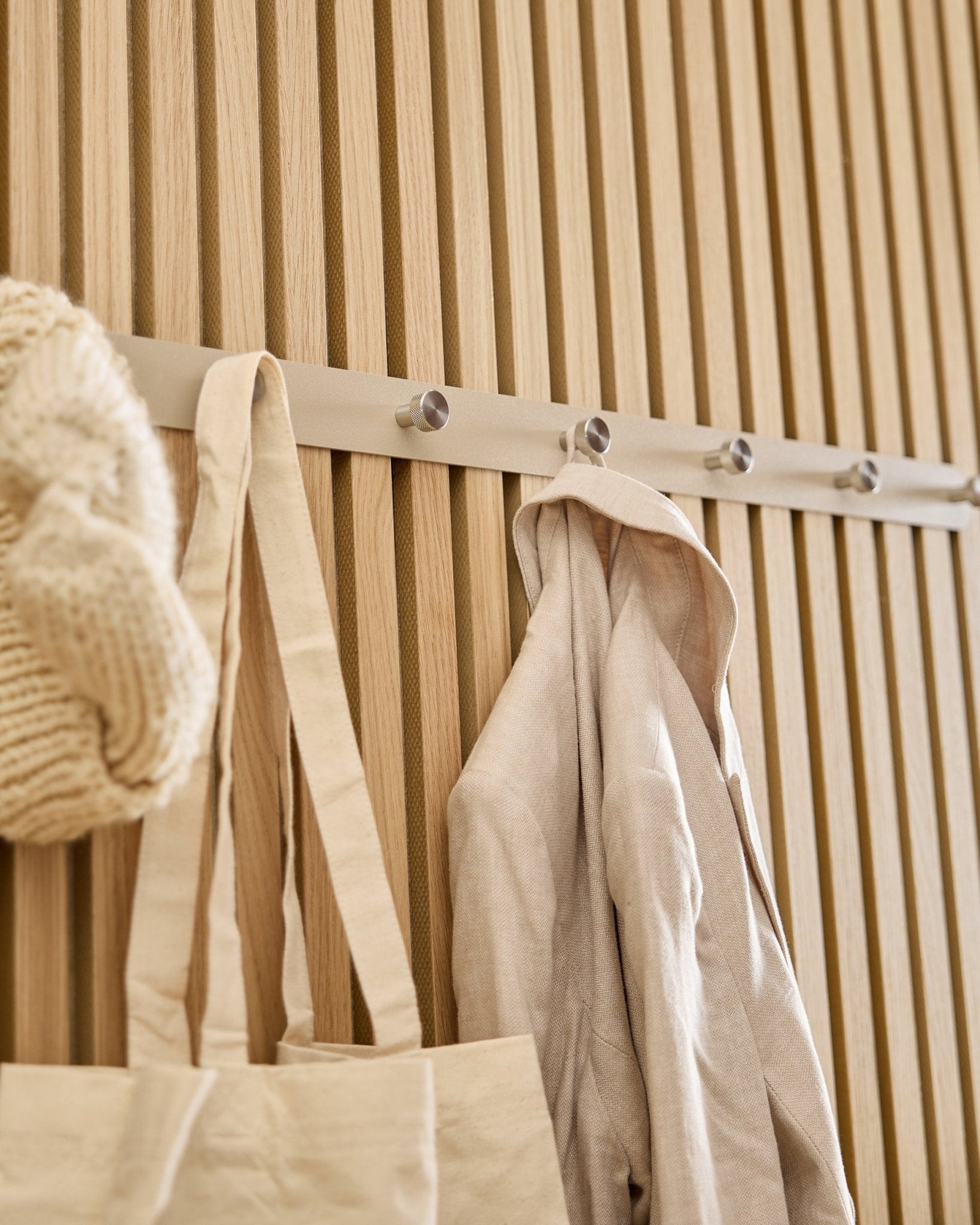 Una tote bag beige, un berretto in maglia e una camicia di lino sono appesi all’appendiabiti a ganci WoodUpp Norr (Beige/Acciaio inox Steel, X7) montato su una slatwall Akupanel in legno chiaro.