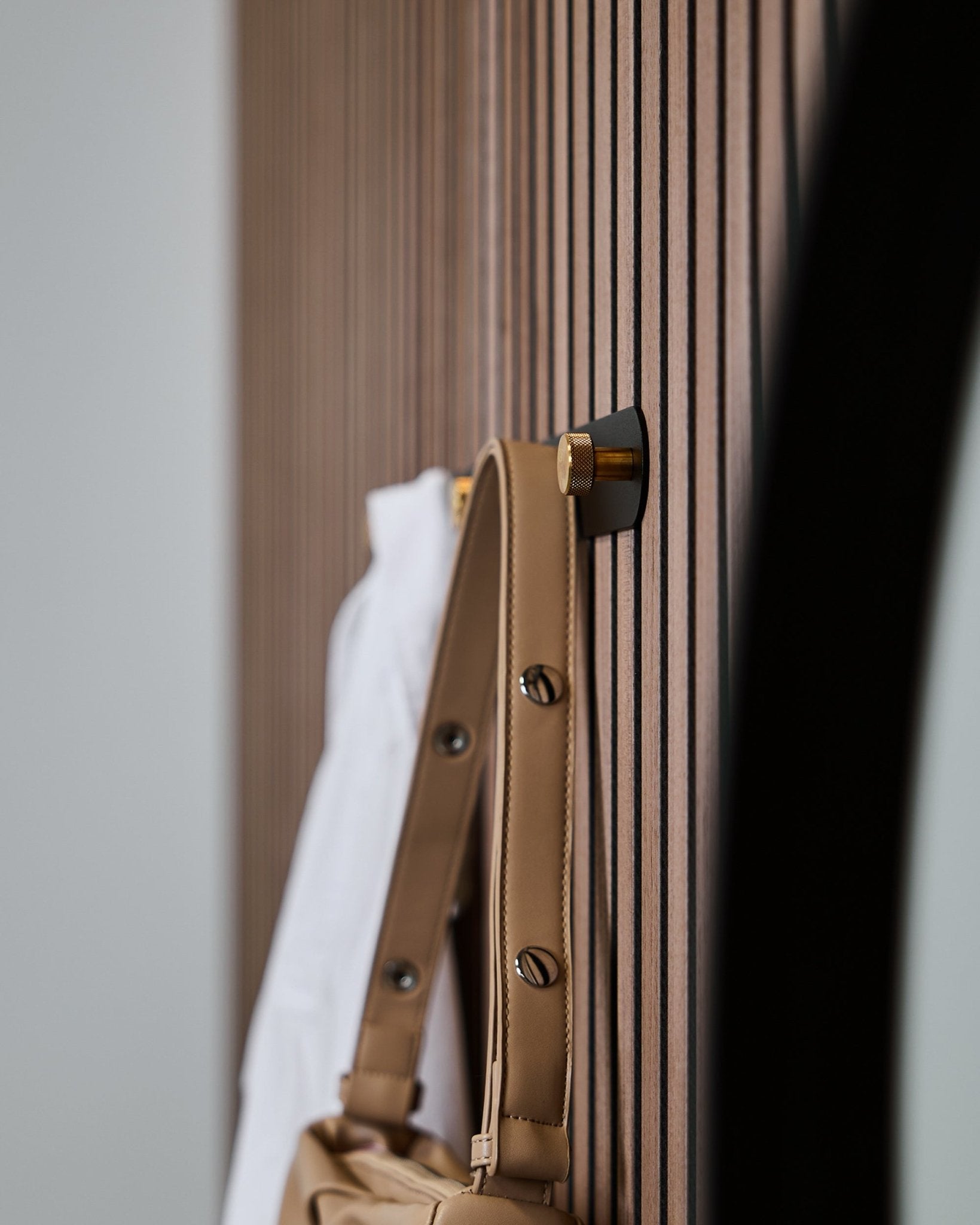 Una borsa color cuoio e una camicia bianca sono appese a un appendiabiti WoodUpp Norr (Black/Brass, X5) montato su un pannello acustico a doghe vertical Akupanel.