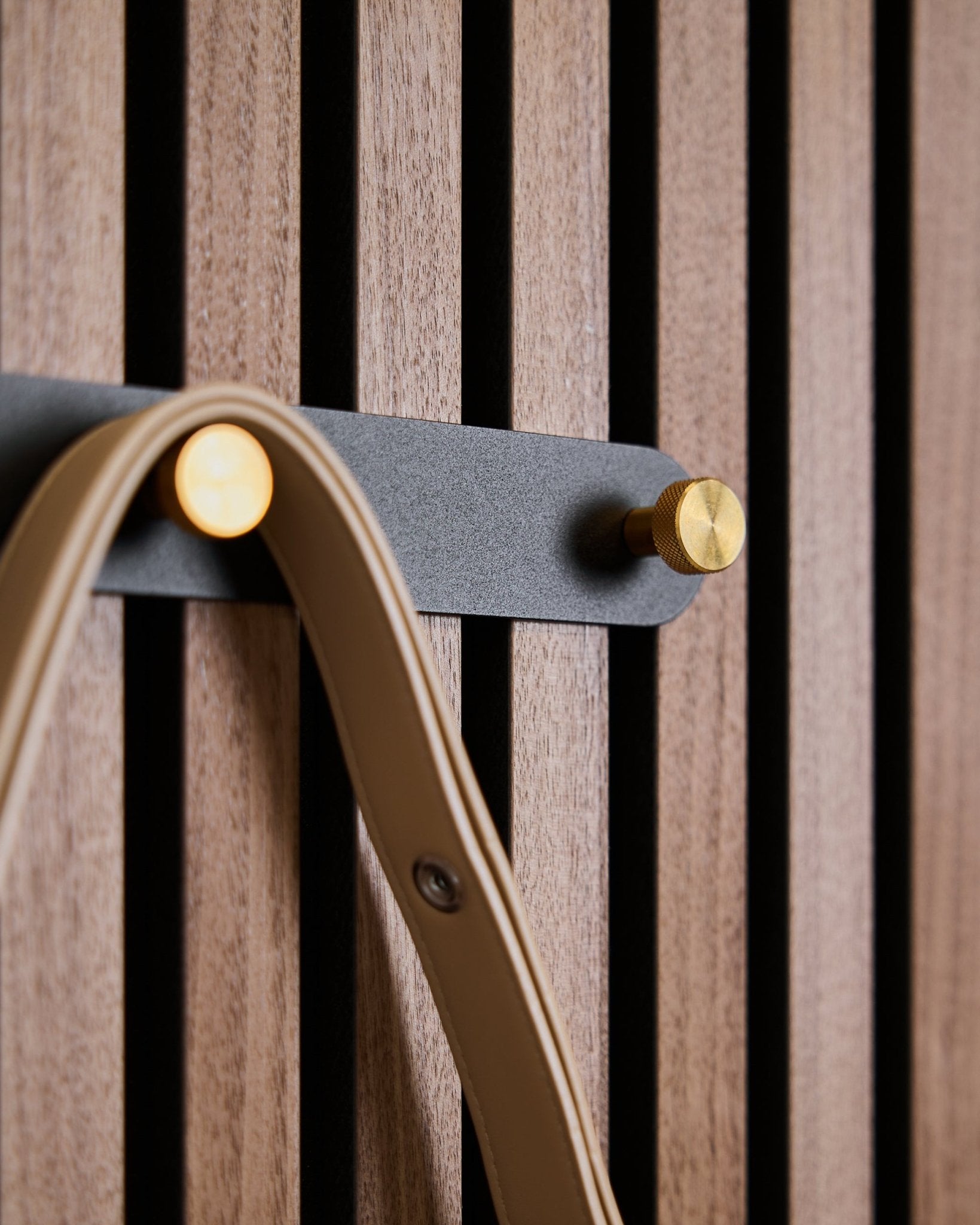 Cinque ganci black/brass dell’appendiabiti Norr di WoodUpp sono montati su un pannello acustico a doghe in legno, ognuno con un laccetto beige appeso.