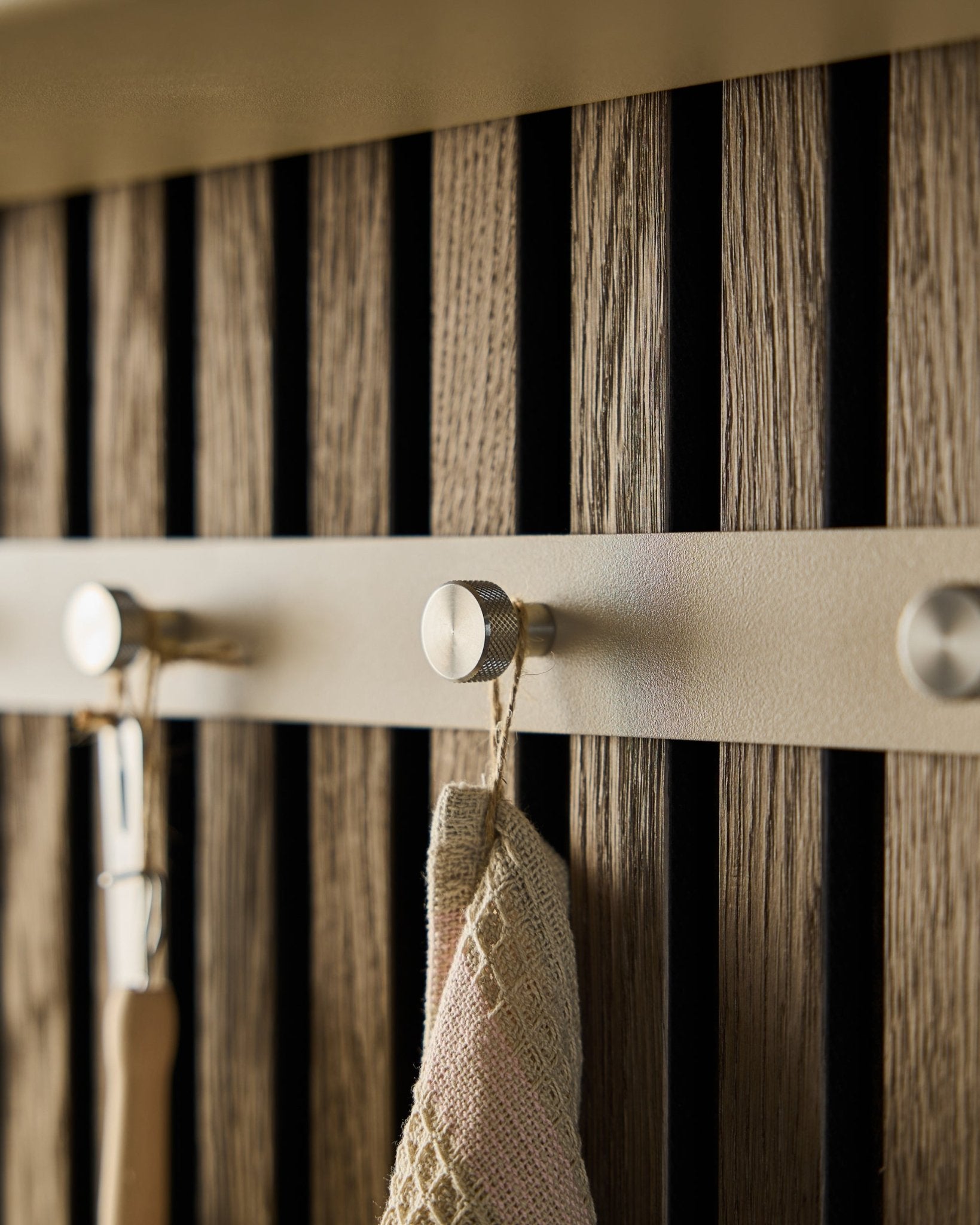 Primo piano dell'appendiabiti WoodUpp Norr Coat Hook Rack (Beige/Steel inox, X5) montato su una parete a doghe in legno Pannello acustico, con un asciugamano e un utensile con manico appesi.