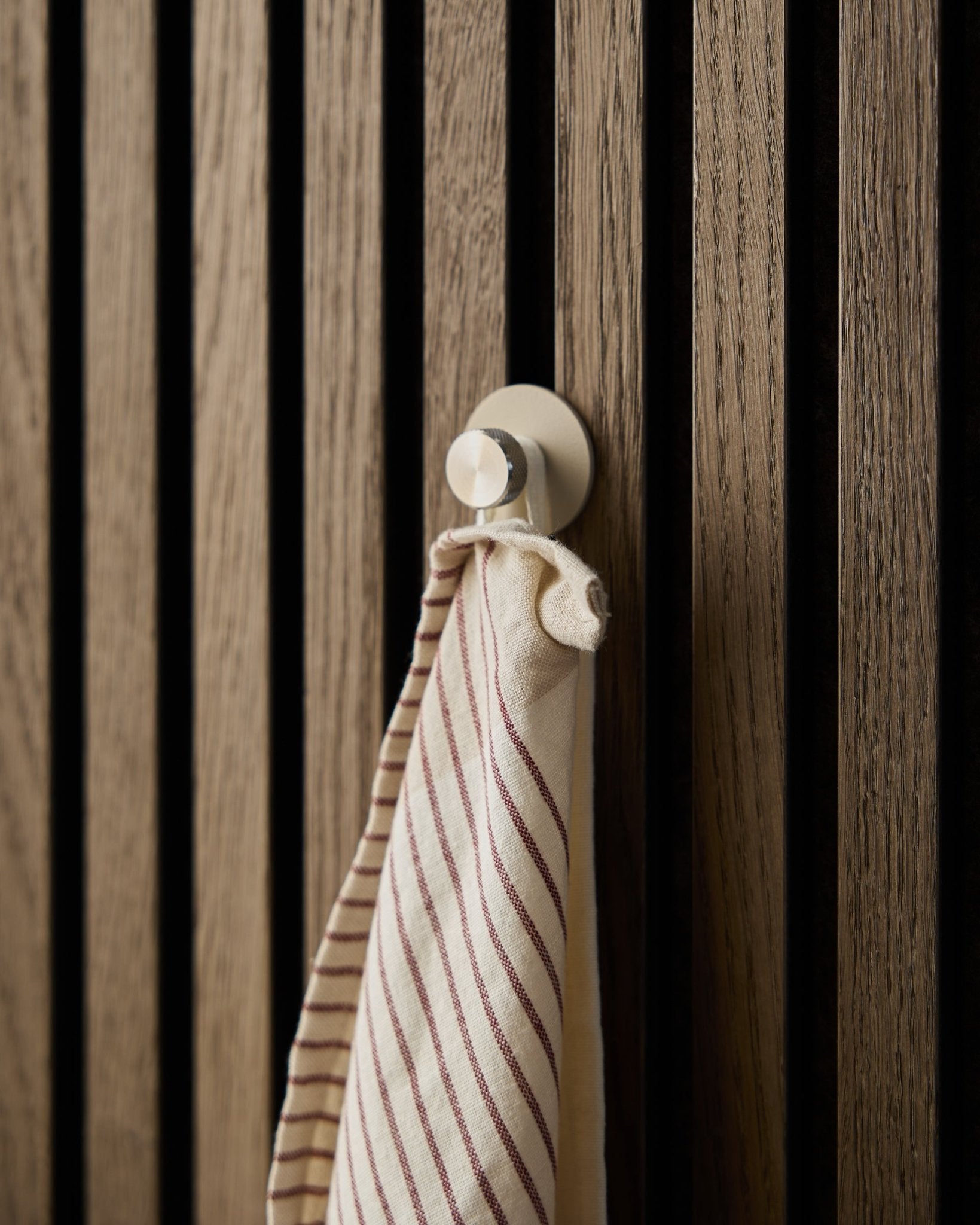 Un asciugamano a righe è appeso a un gancio appendiabiti beige Norr di WoodUpp, realizzato in acciaio inox steel, montato su un pannello acustico a doghe in legno Akupanel dalla texture materica.