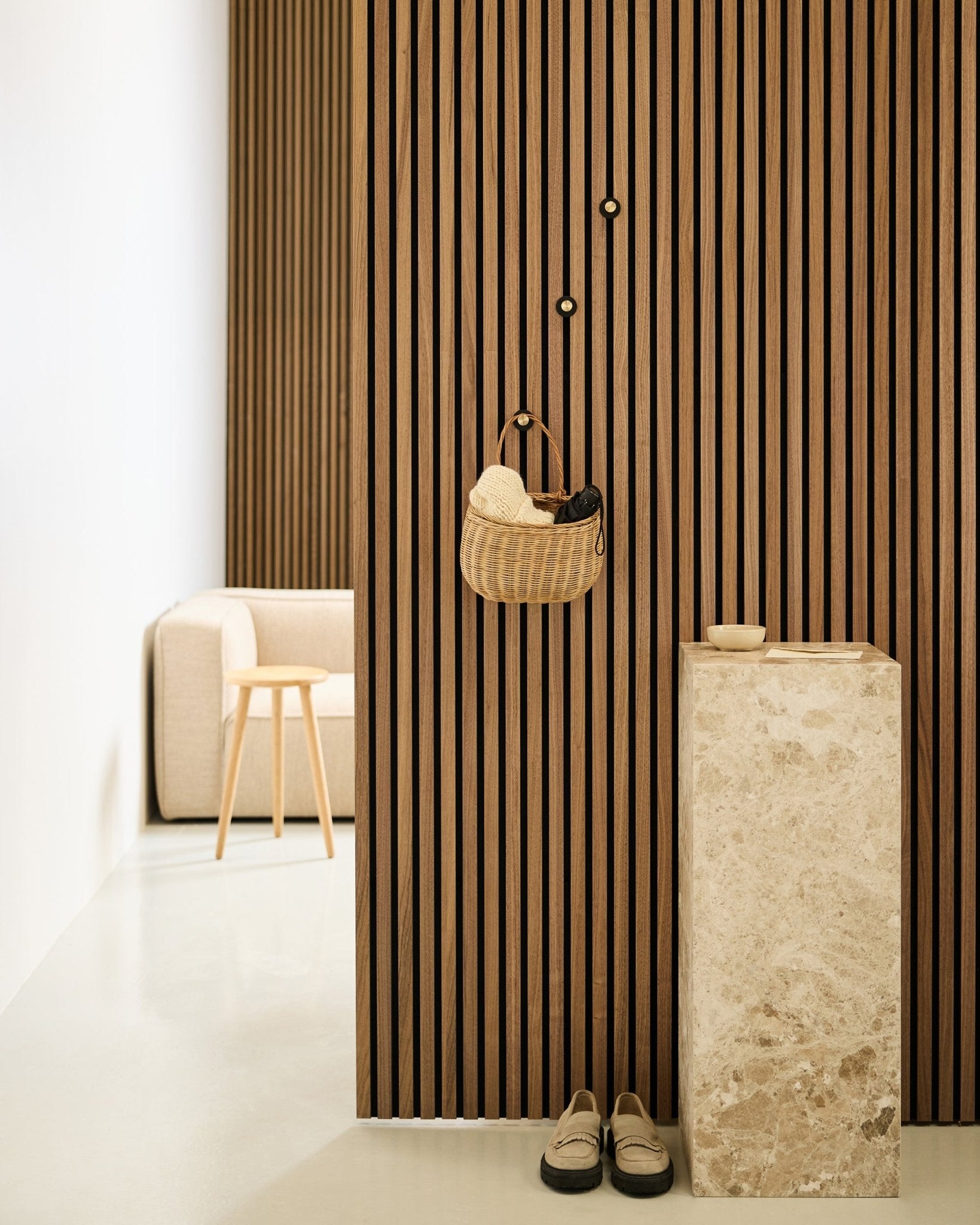 Ingresso minimalista con parete a listelli acustici Akupanel, gancio appendiabiti WoodUpp Norr in black e brass, cesta, piedistallo in pietra, pantofole e sedia beige.