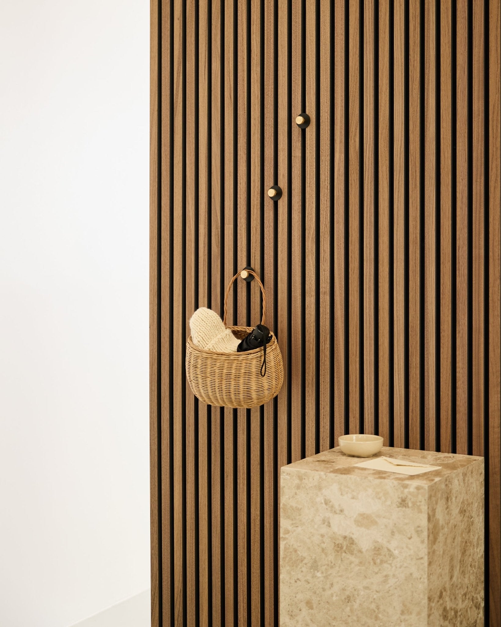Gancio appendiabiti Norr di WoodUpp montato su una parete a listelli acustici nordica Akupanel, accanto a un piedistallo in pietra beige con una ciotola small in un interno minimalista.