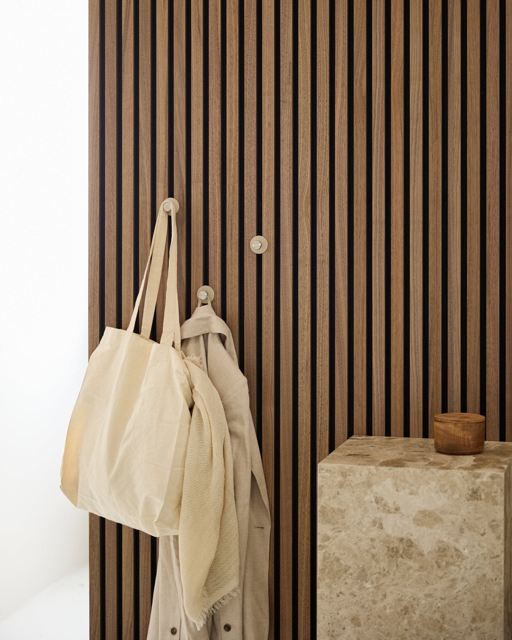Una tote bag beige e un tessuto leggero sono appesi a un gancio appendiabiti WoodUpp Norr (Beige/acciaio inox Steel), montato su un vertical Akupanel Pannello acustico accanto a una superficie in pietra.