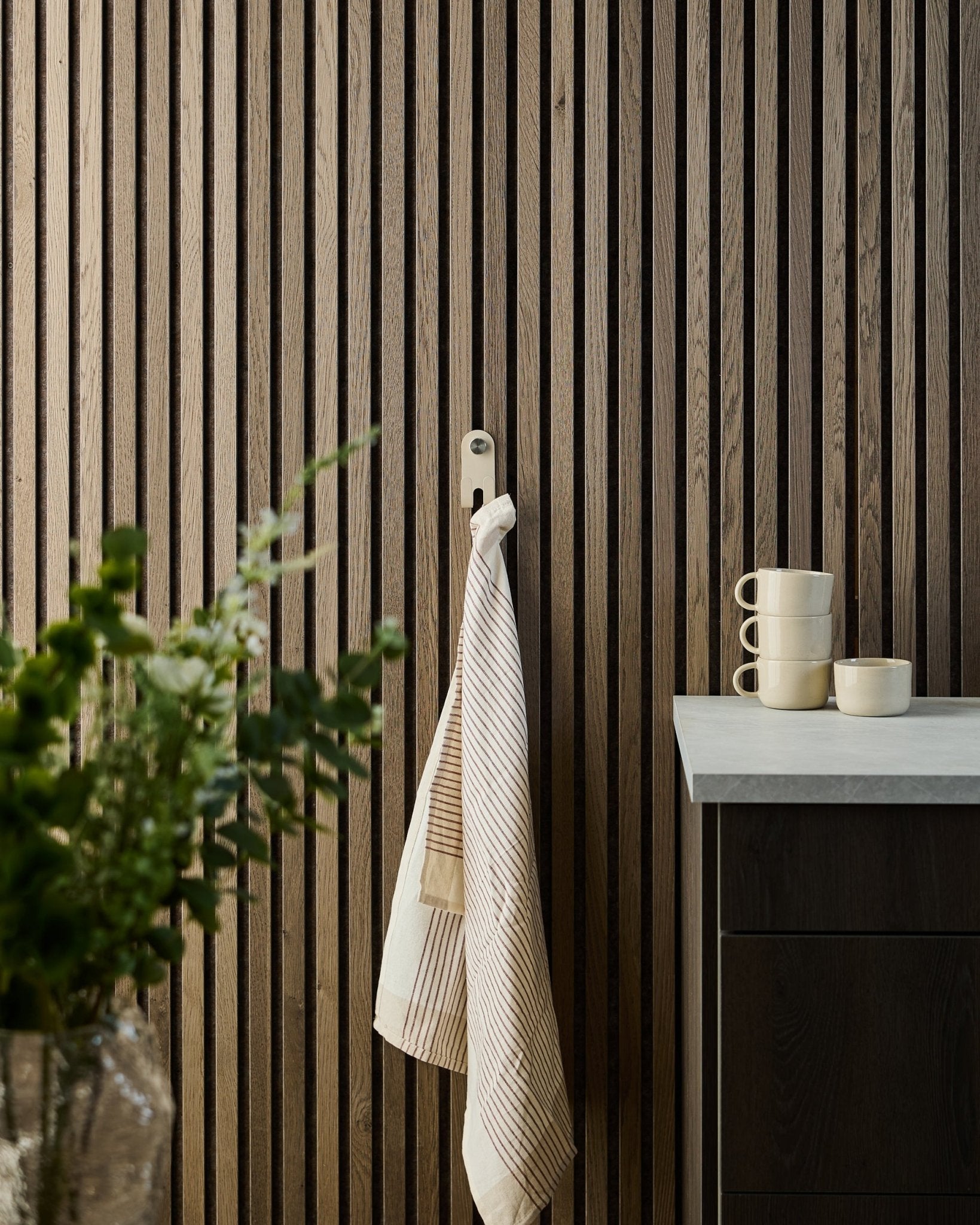 Uno strofinaccio a righe è appeso al WoodUpp Norr Double Hanger in beige, montato su un pannello fonoassorbente a doghe nordico; sotto si vedono tazze, una ciotola e una pianta verde.