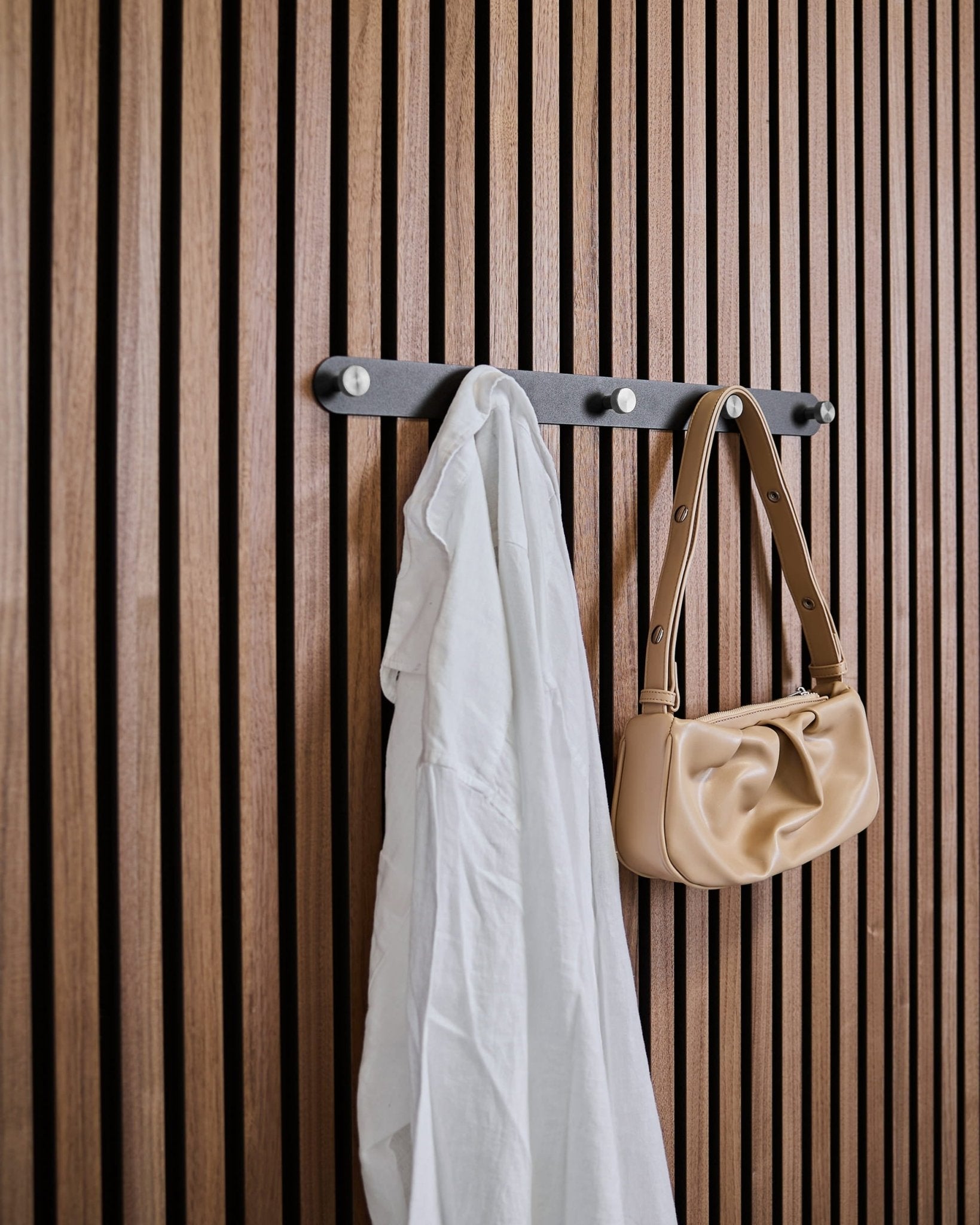Una camicia bianca e una borsa beige sono appese ai ganci appendiabiti WoodUpp Norr X5 Black/Stainless Steel, fissati a un pannello acustico a doghe vertical.
