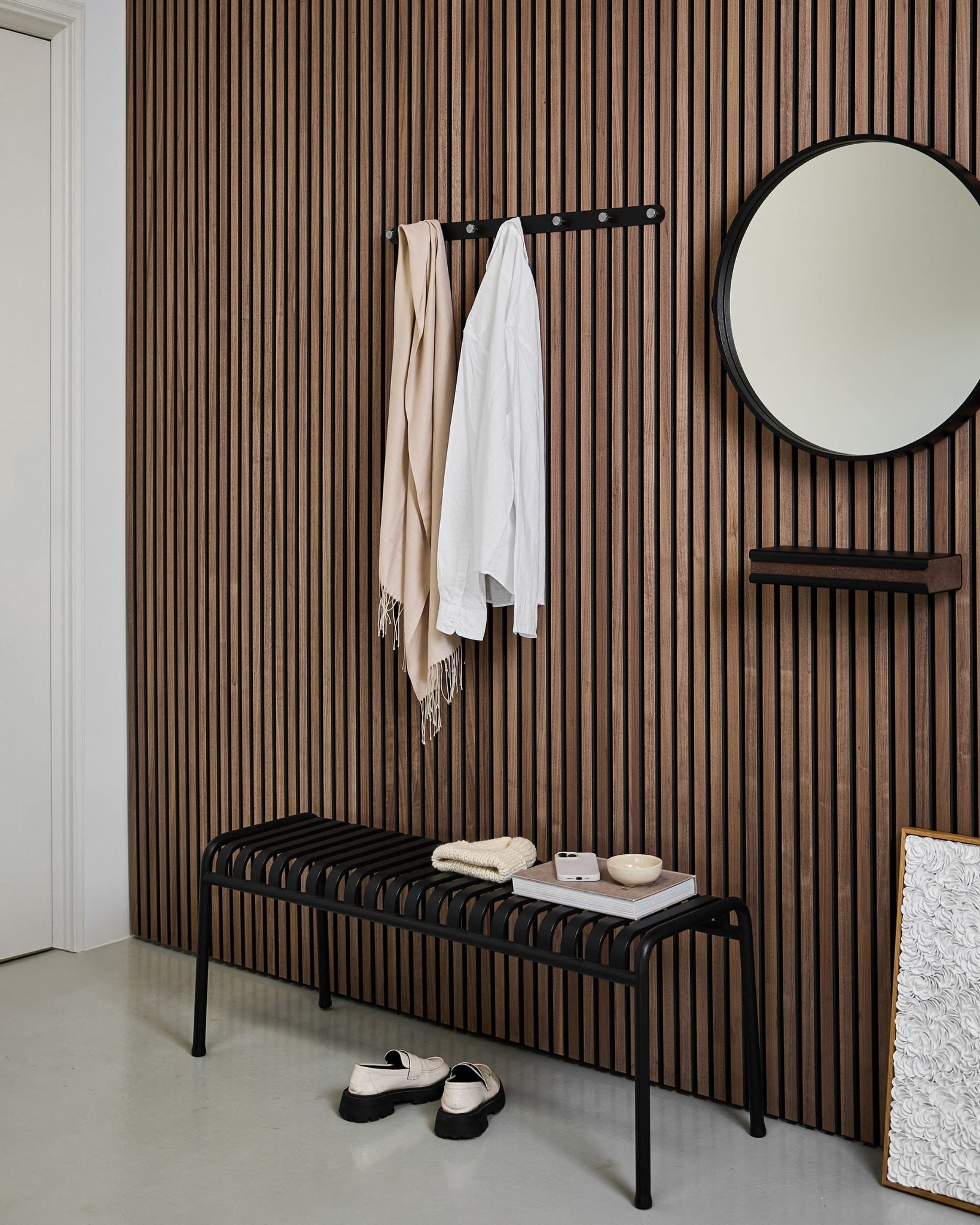 Ingresso moderno con parete in pannelli acustici in legno a doghe in stile nordico, specchio rotondo, panca black, appendiabiti a ganci WoodUpp Norr e pantofole su un pavimento chiaro.