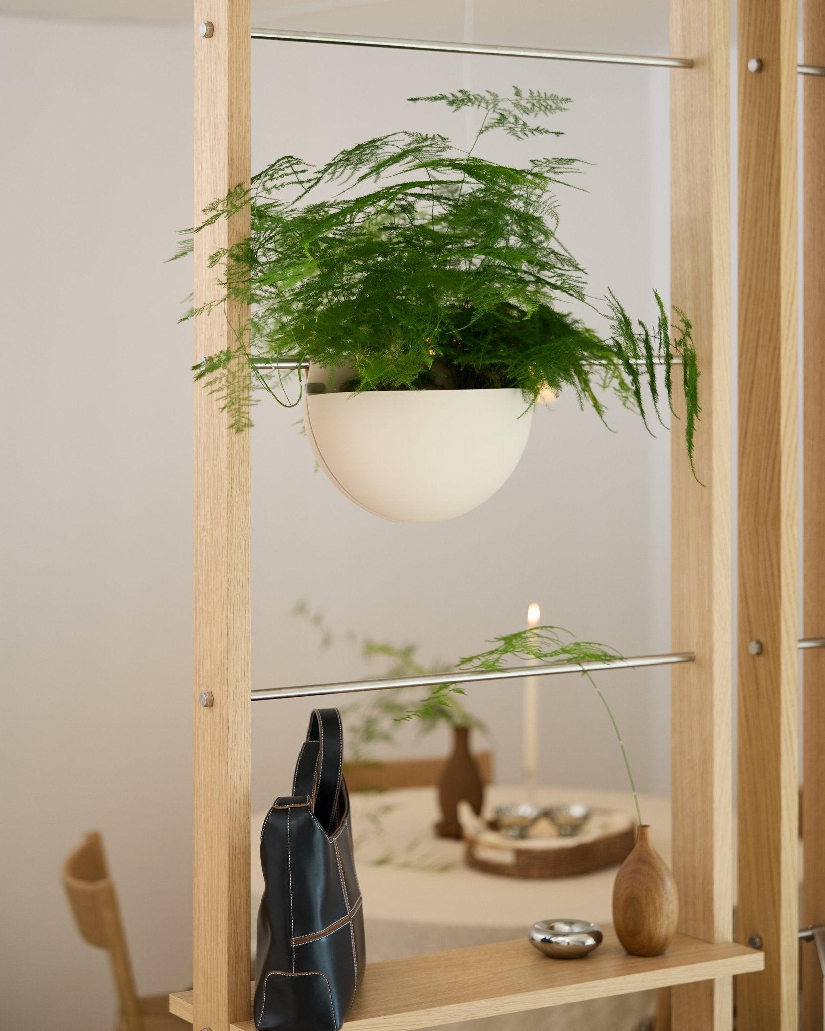 Una felce verde, collocata in una fioriera WoodUpp Gro Soft Pocket BM2 color beige chiaro, poggia su una mensola di legno sopra una borsa nera, un vaso e una tavola imbandita.