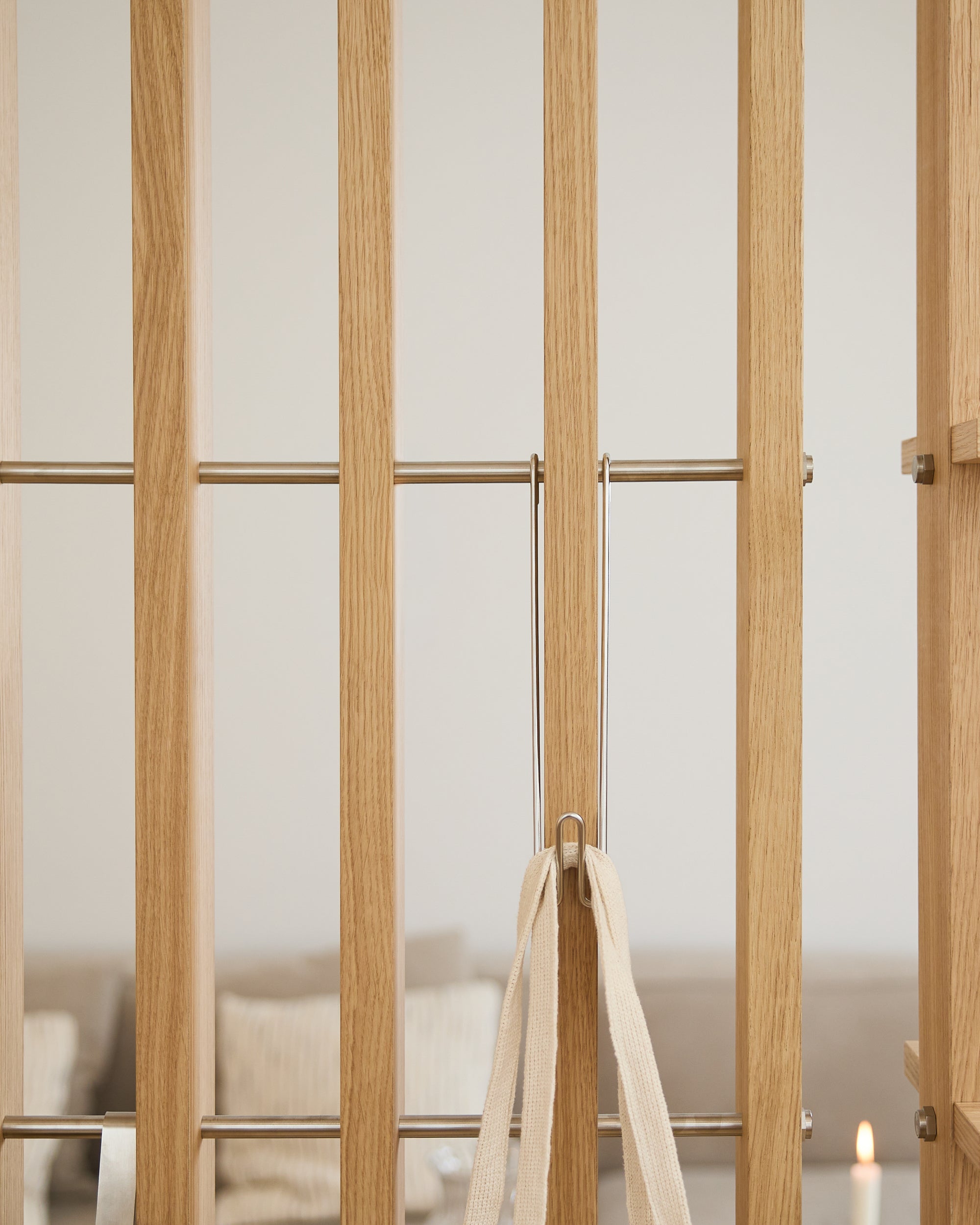 Cancello in legno chiaro con barre metalliche, nastro beige e tre ganci WoodUpp Alva Wire Hooks/BM1; divano e candela sfocati sullo sfondo.