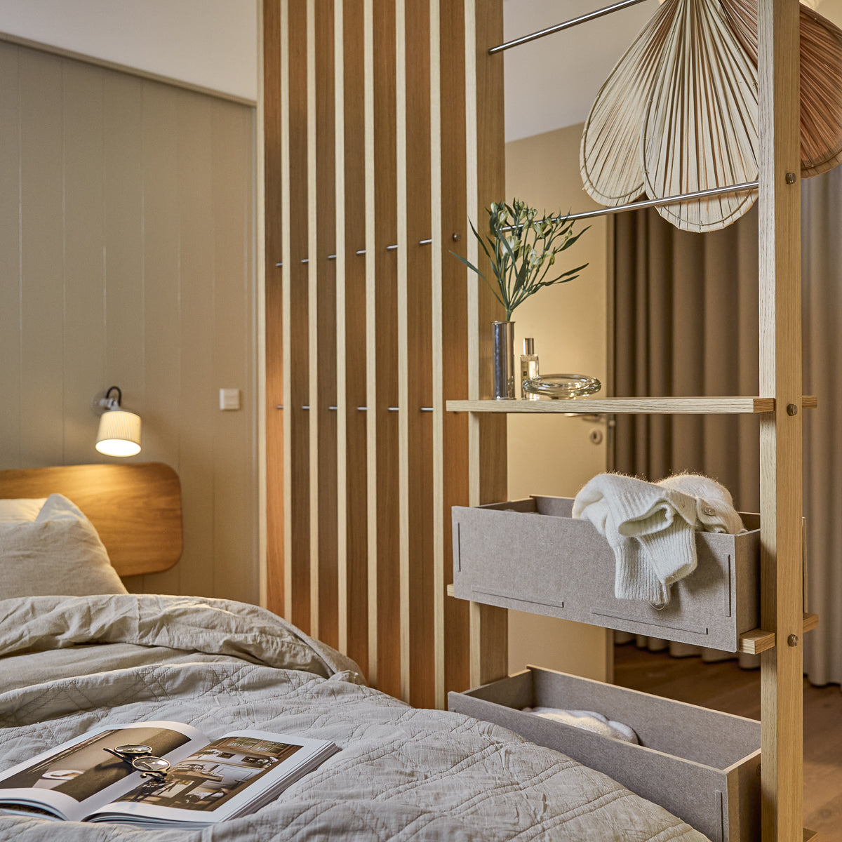 Una camera da letto moderna con biancheria da letto morbida e nordica, una lampada a sospensione e una rivista sul letto ben sistemato. I pannelli acustici in legno WoodUpp Akupanel accentuano la parete.