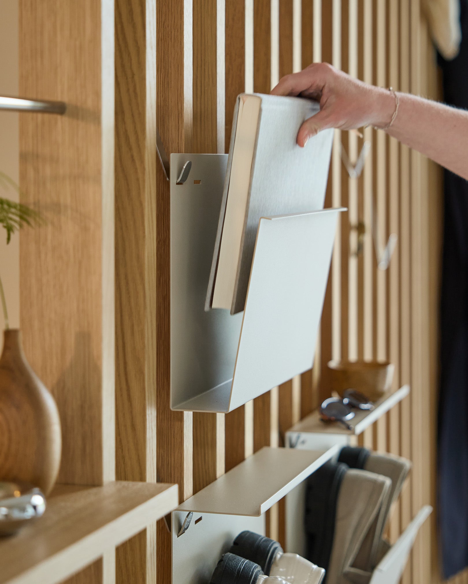 Una mano appoggia Nell | Magazine Storage / Beige di WoodUpp su una mensola in steel verniciata a polvere, montata su un elemento nordico in legno Pannello acustico, con altri oggetti nelle vicinanze.