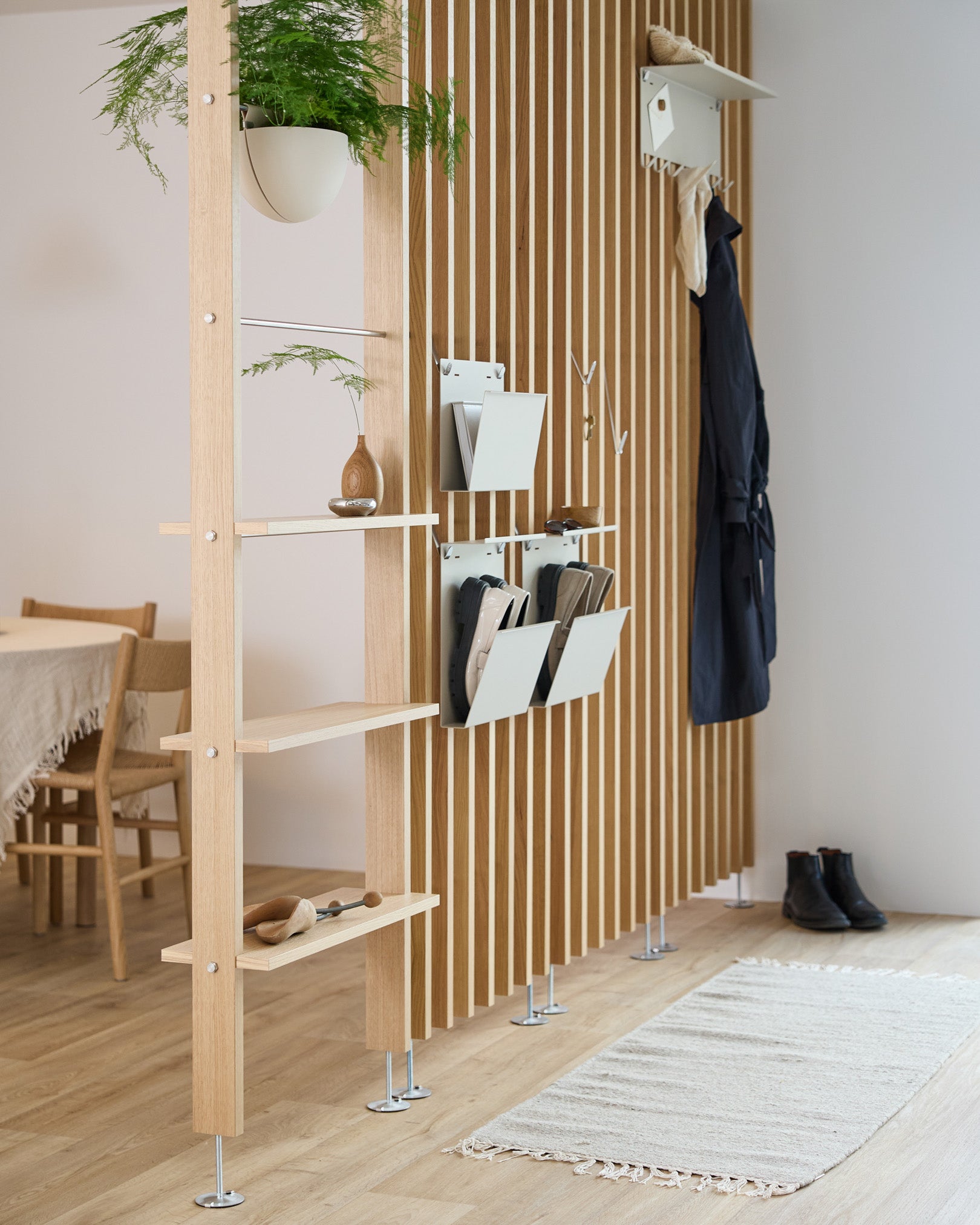 Il portariviste Nell di WoodUpp in beige è integrato in un divisorio per ambienti con mensole in steel, contenitori portaoggetti e ganci appendiabiti; sullo sfondo si vedono la zona pranzo e un tappeto.
