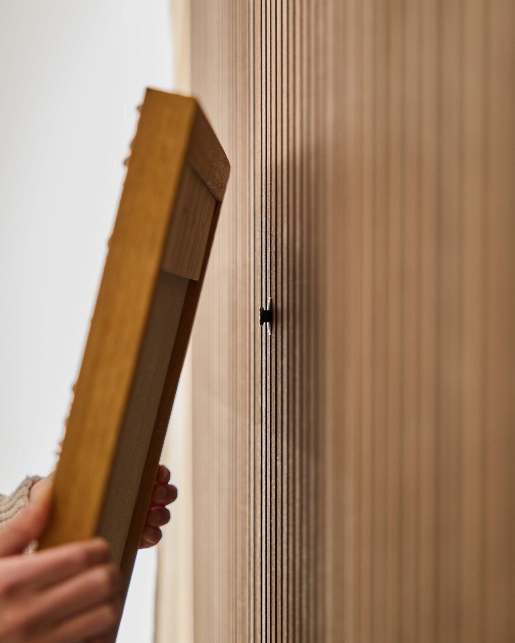 Una persona usa il set WoodUpp Dupps | Original per montare una cornice per pannello acustico in legno Akupanel, concentrandosi sul fissaggio di un gancio small black vicino alle doghe vertical.