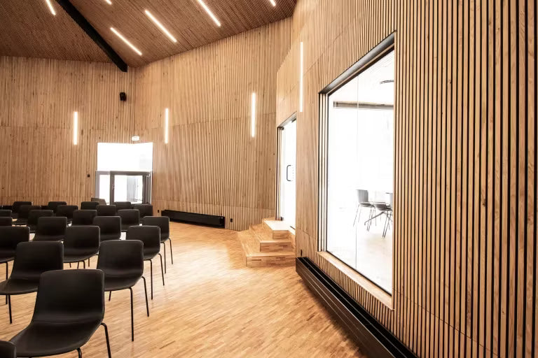 Ampia sala conferenze con pannelli acustici in legno Akupanel alle pareti e al soffitto, file di sedie nere, ampie finestre e apparecchi di illuminazione verticali.