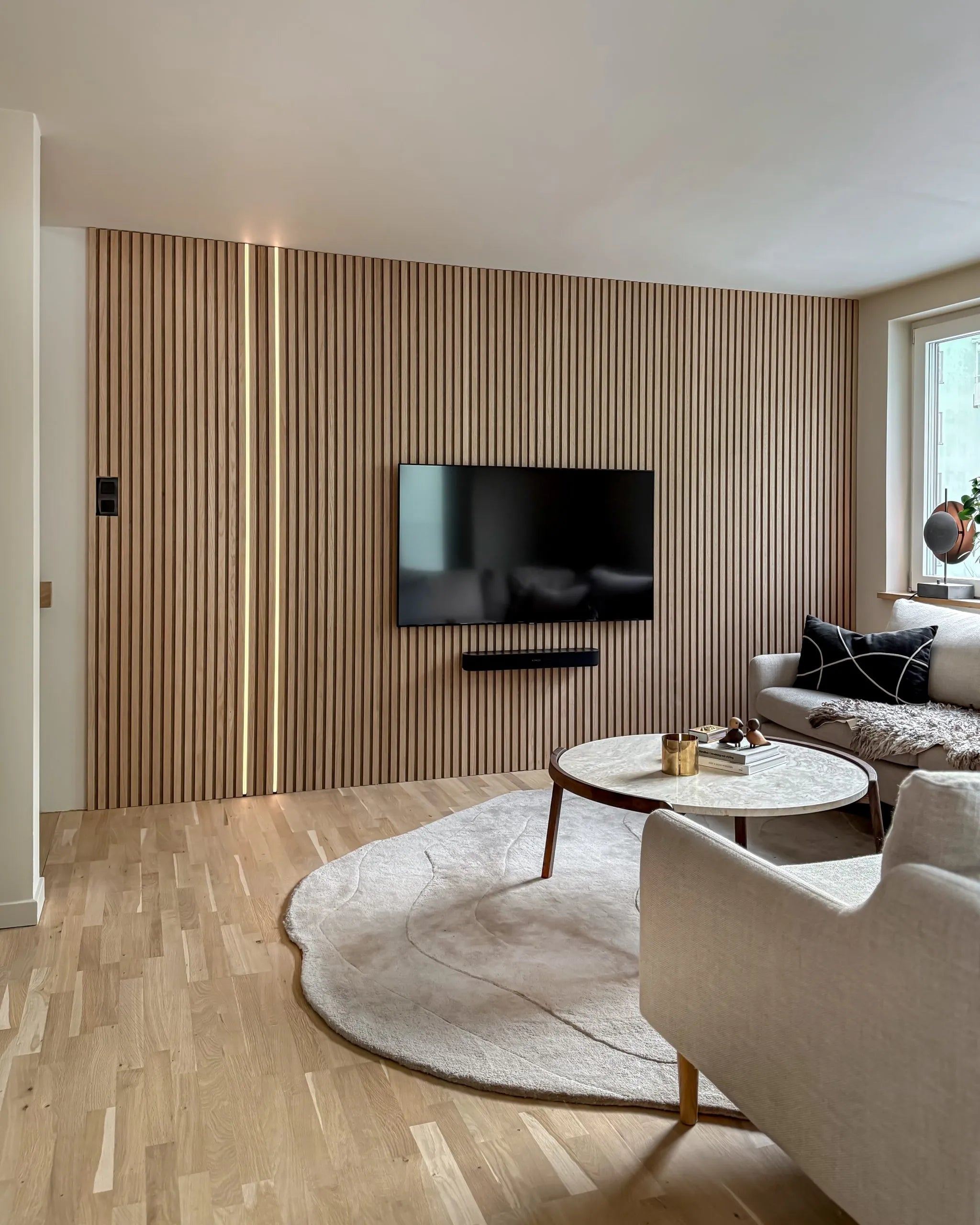 Soggiorno moderno con parete daccento in doghe di legno acustico nordico Akupanel, TV montata, tavolino rotondo, divano color crema, grande tappeto su pavimenti in legno chiaro.