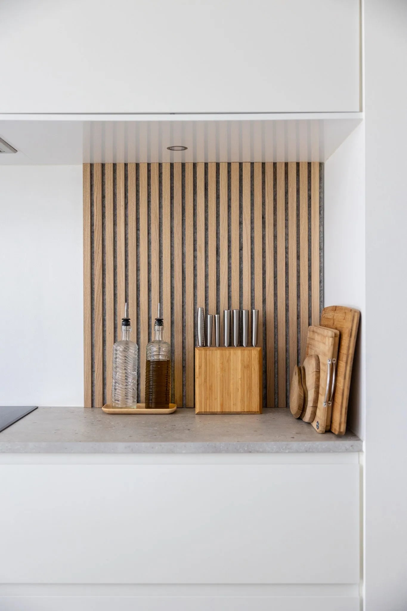 Bancone da cucina moderno con bottiglie dolio, ceppo di coltelli in legno e taglieri impilati su un backsplash a doghe in legno acustico Akupanel nordico e armadietti bianchi.