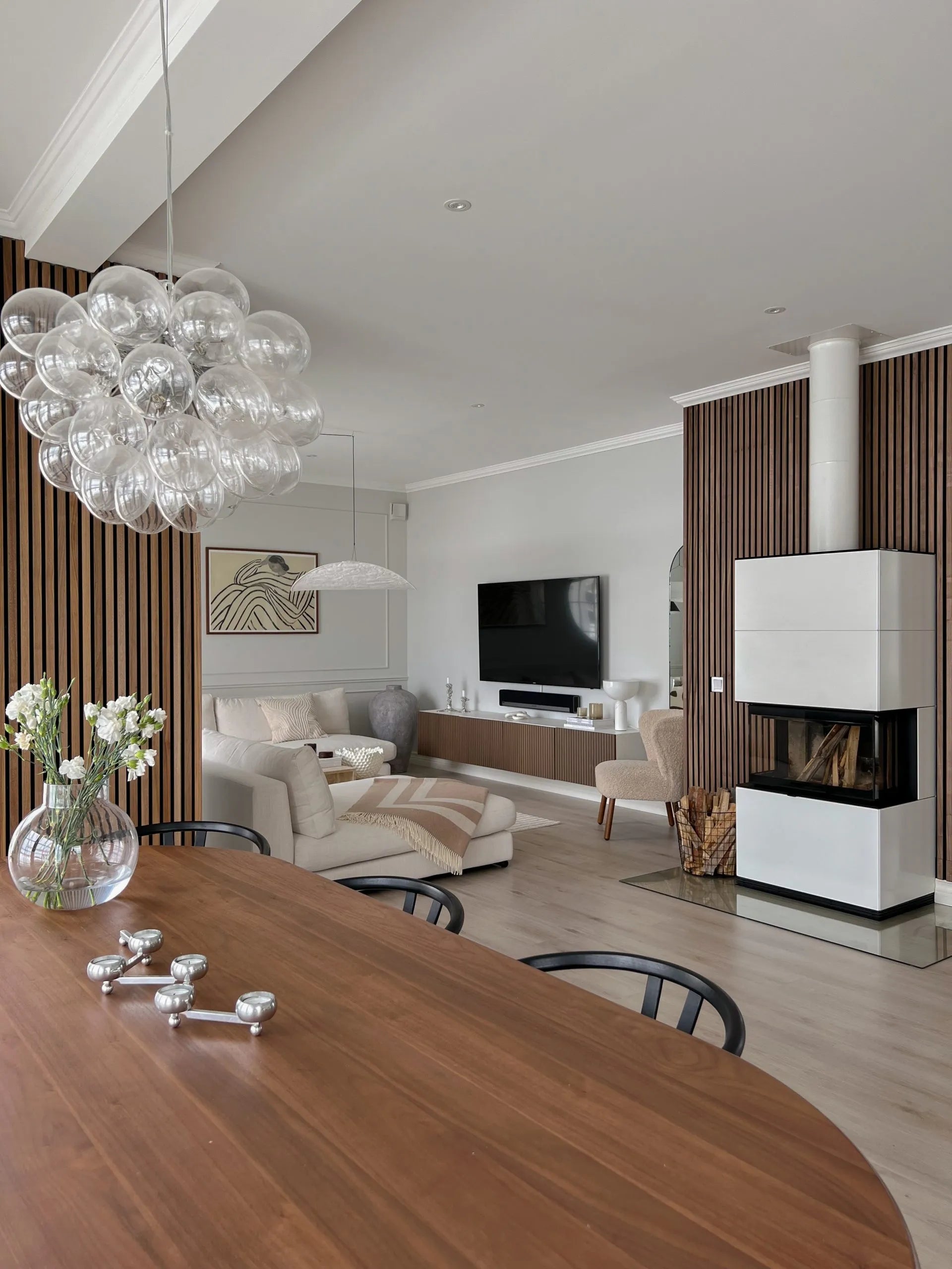Moderna zona giorno e pranzo con pannelli acustici in legno nordico, comodi posti a sedere, TV, elegante caminetto con legna accatastata e lampadario a bolle di vetro.