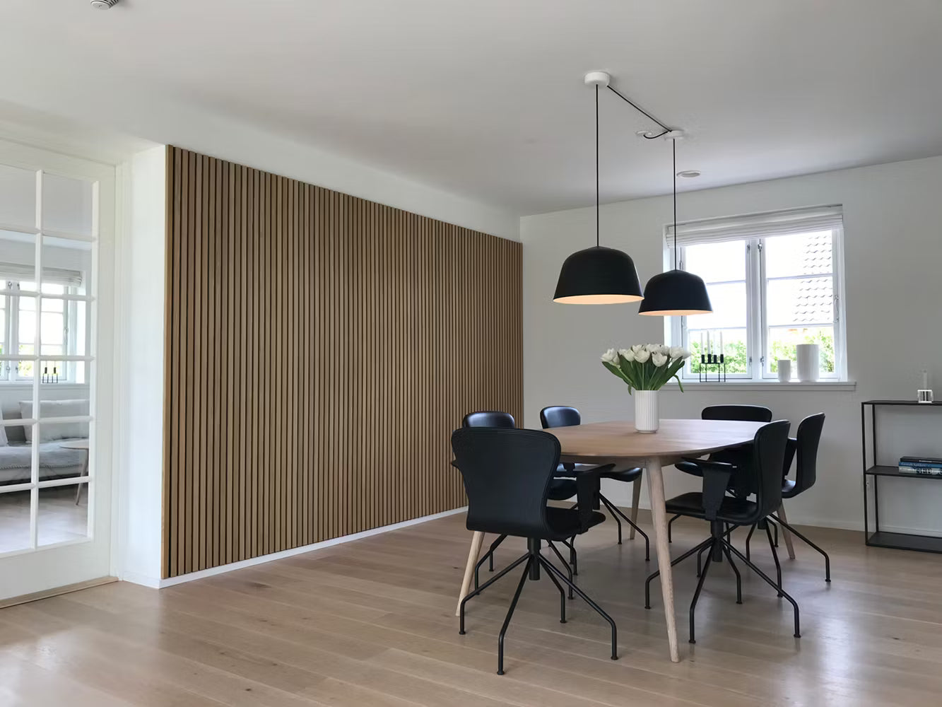 Sala da pranzo moderna con pavimento in legno, parete acustica a doghe Akupanel, sedie nere, tavolo in legno, luci a sospensione e centrotavola floreale bianco.
